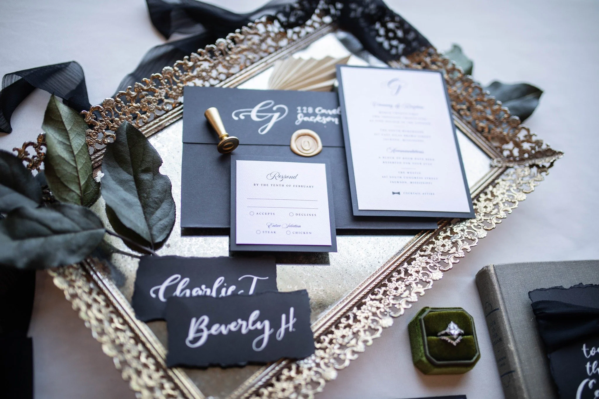 Wedding invitation suite on a decorative mirror with black foliage, a gold and black ring box, and place cards for Charlie T and Beverly H.