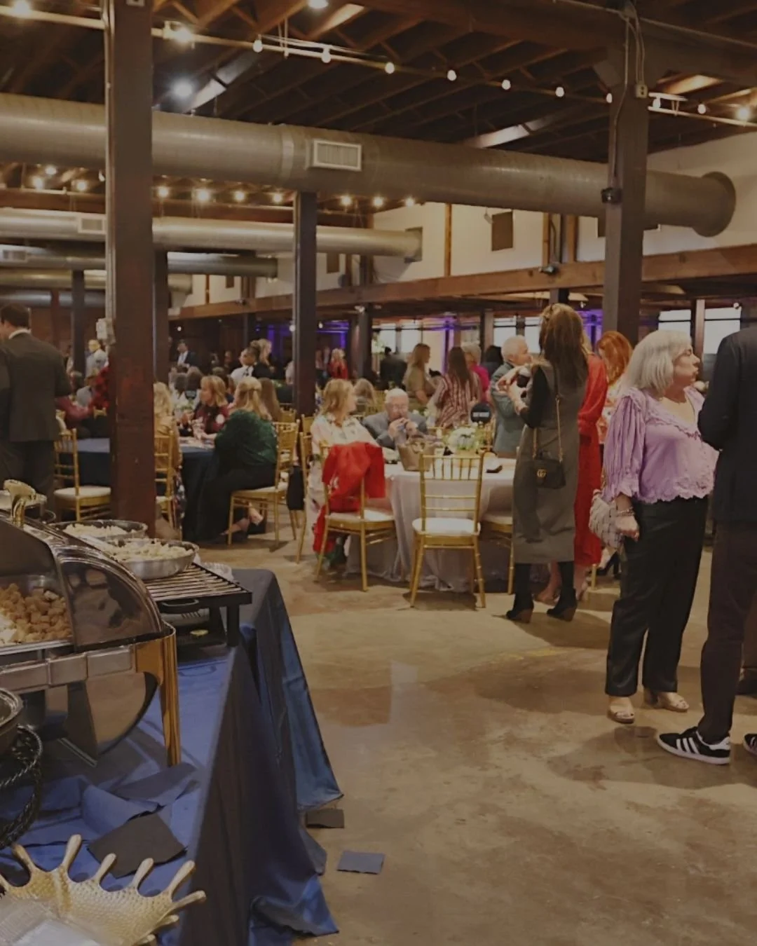 Indoor banquet hall filled with guests sitting at tables and standing in groups, with buffet food on a table in the foreground, wooden beams on the ceiling, and string lights hanging overhead.