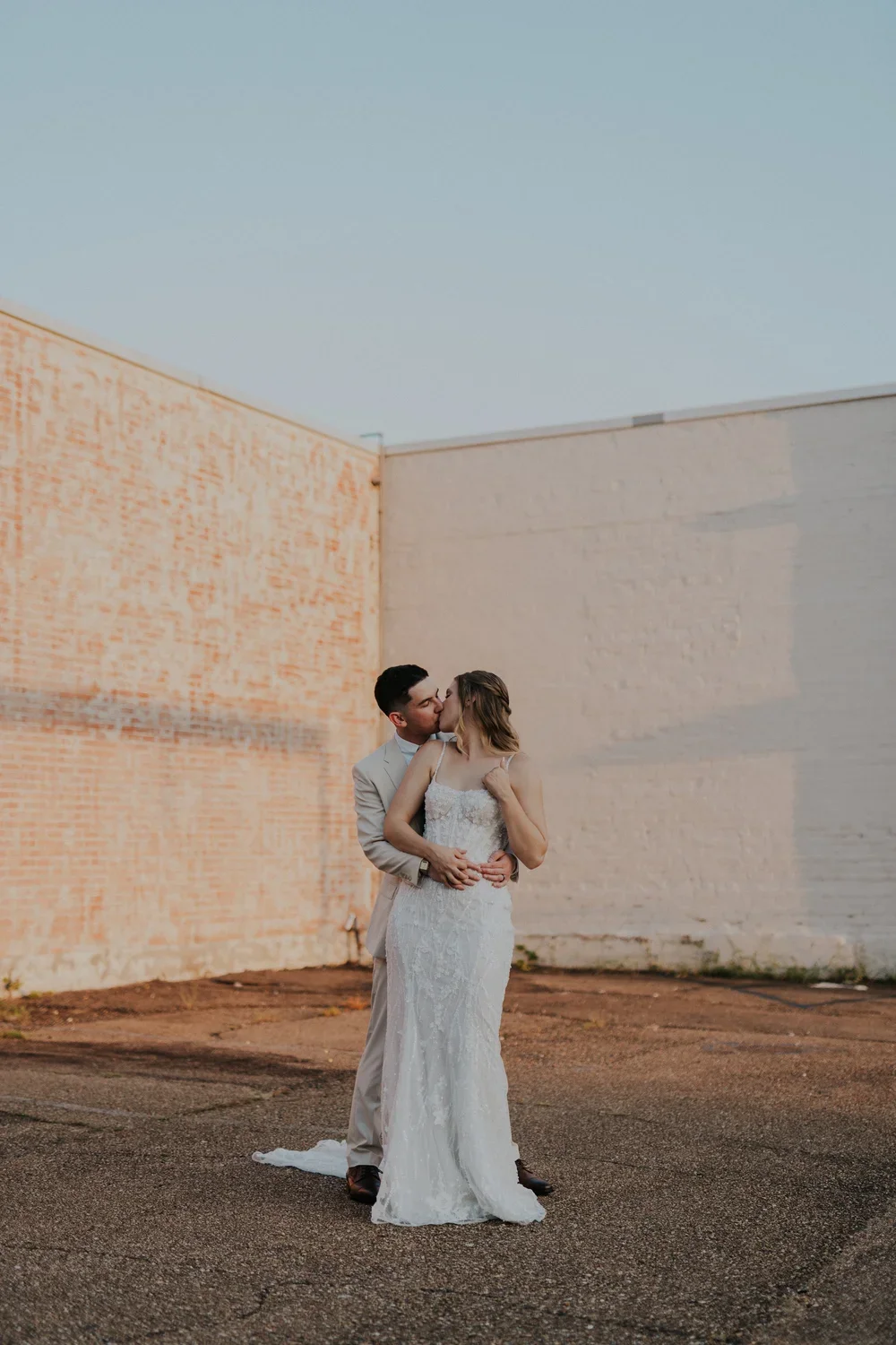 A couple dressed in wedding attire sharing a kiss outdoors against a brick and white wall with a clear sky overhead.