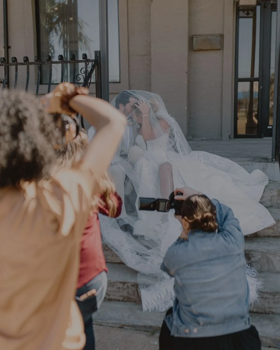 Two photographers taking pictures of a bride and groom sitting on steps outside a building, with the bride wearing a veil.