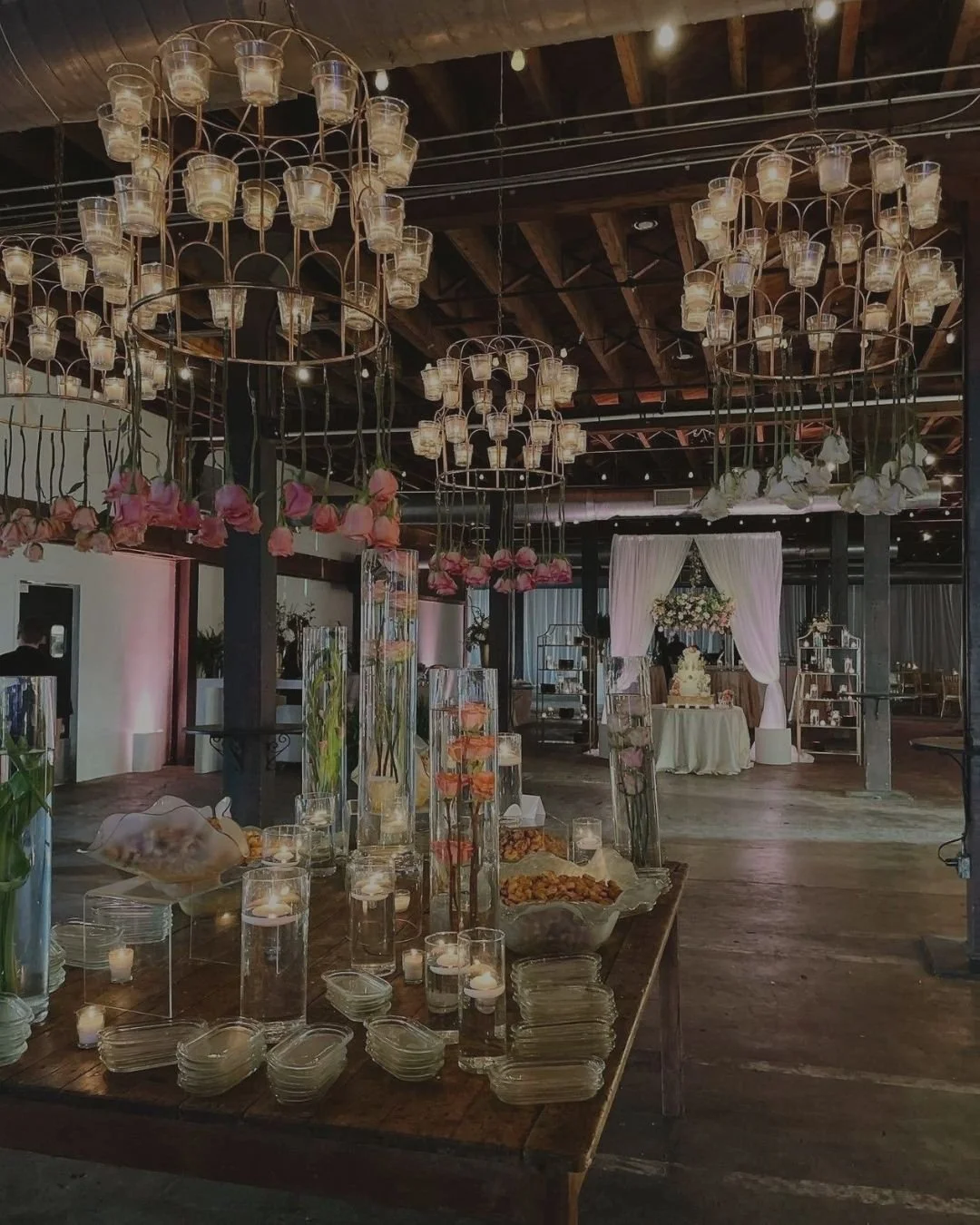 An indoor event space decorated with chandelier-style lighting fixtures and floral arrangements. A table holds candles, glasses, bowls with food, and tall vases with flowers. In the background, a draped fabric backdrop surrounds a cake table.