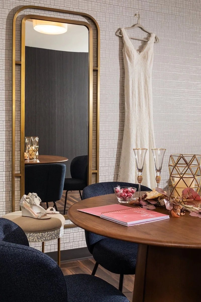 Wedding dress hanging on a wall mirror in a modern interior with a round wooden table decorated with pink flowers, gold candlesticks, a pink folder, and a pair of white shoes, with a mirror reflecting chairs and part of a ceiling light.