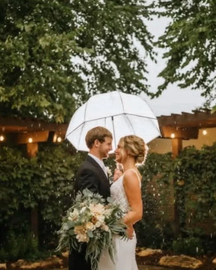A couple in wedding attire sharing a romantic moment under an umbrella outdoors, surrounded by greenery and string lights.