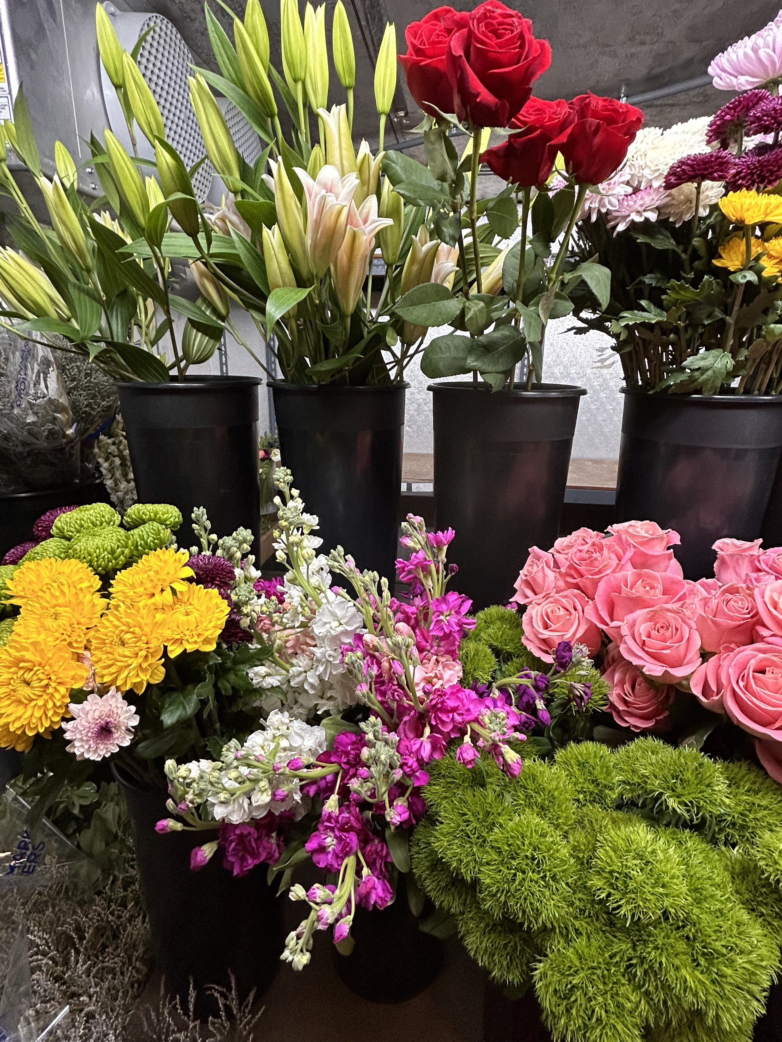 Various colorful flowers including roses, lilies, chrysanthemums, and statice arranged in black pots at a flower shop.