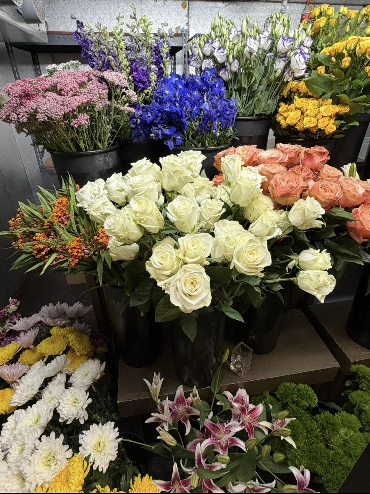 Various colorful flower arrangements including white roses, pink roses, purple irises, yellow roses, blue delphinium, and mixed flowers displayed in black pots at a flower shop.