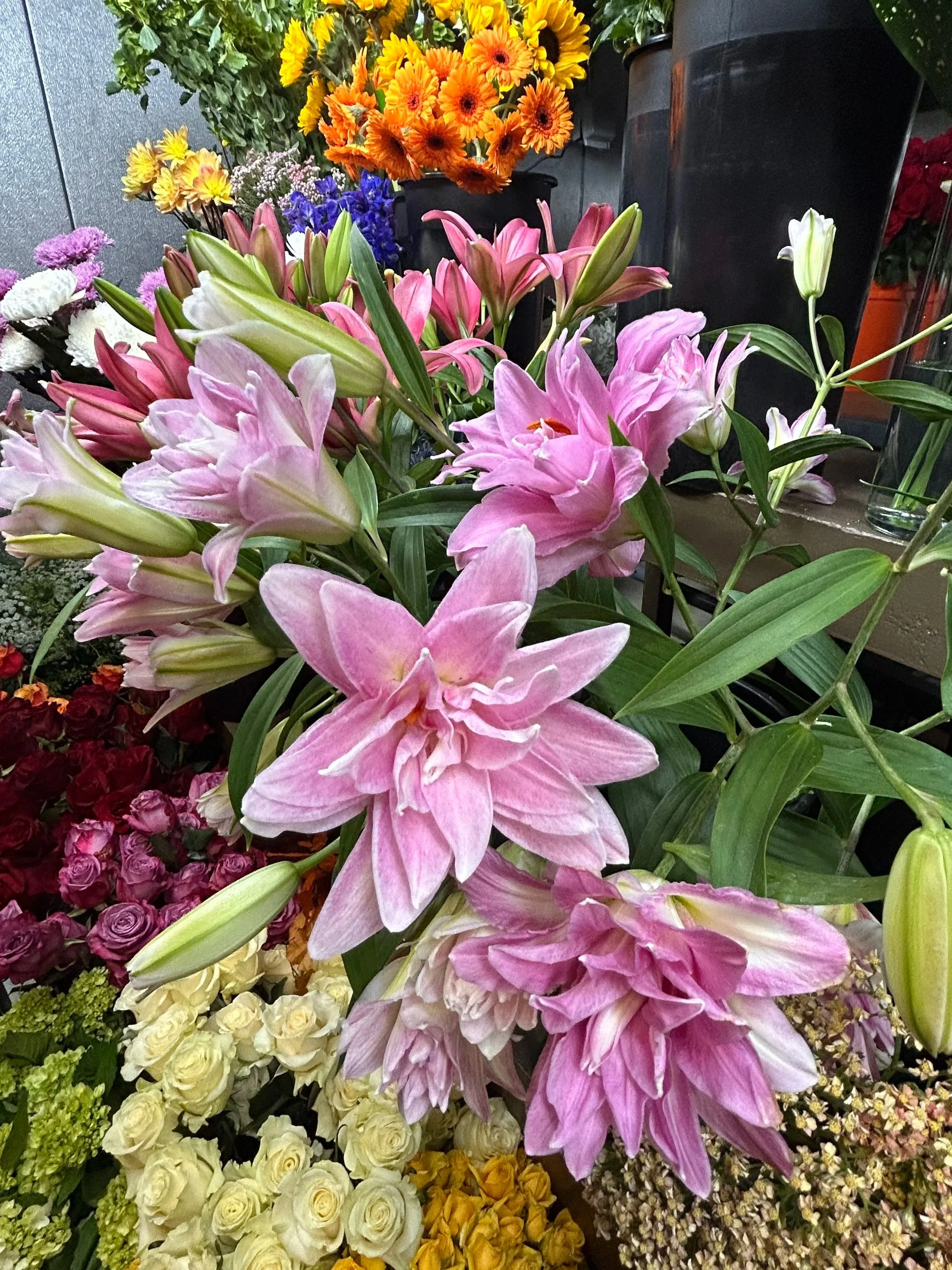 A vibrant display of various flowers, primarily featuring pink lilies and other colorful blossoms in the background in a floral shop setting.