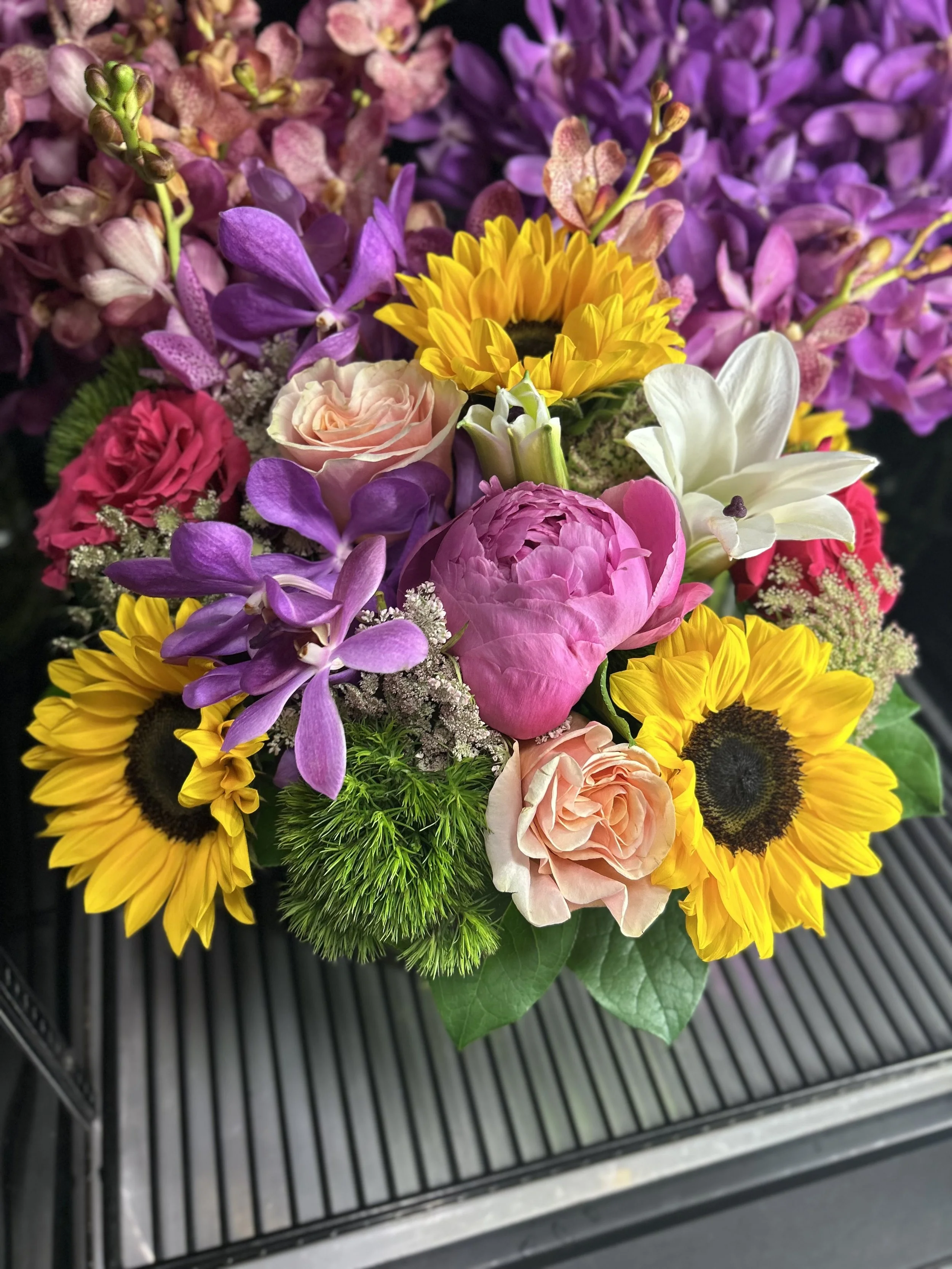 Colorful bouquet of flowers including sunflowers, pink peony, roses, white lily, purple orchids, and assorted greenery.