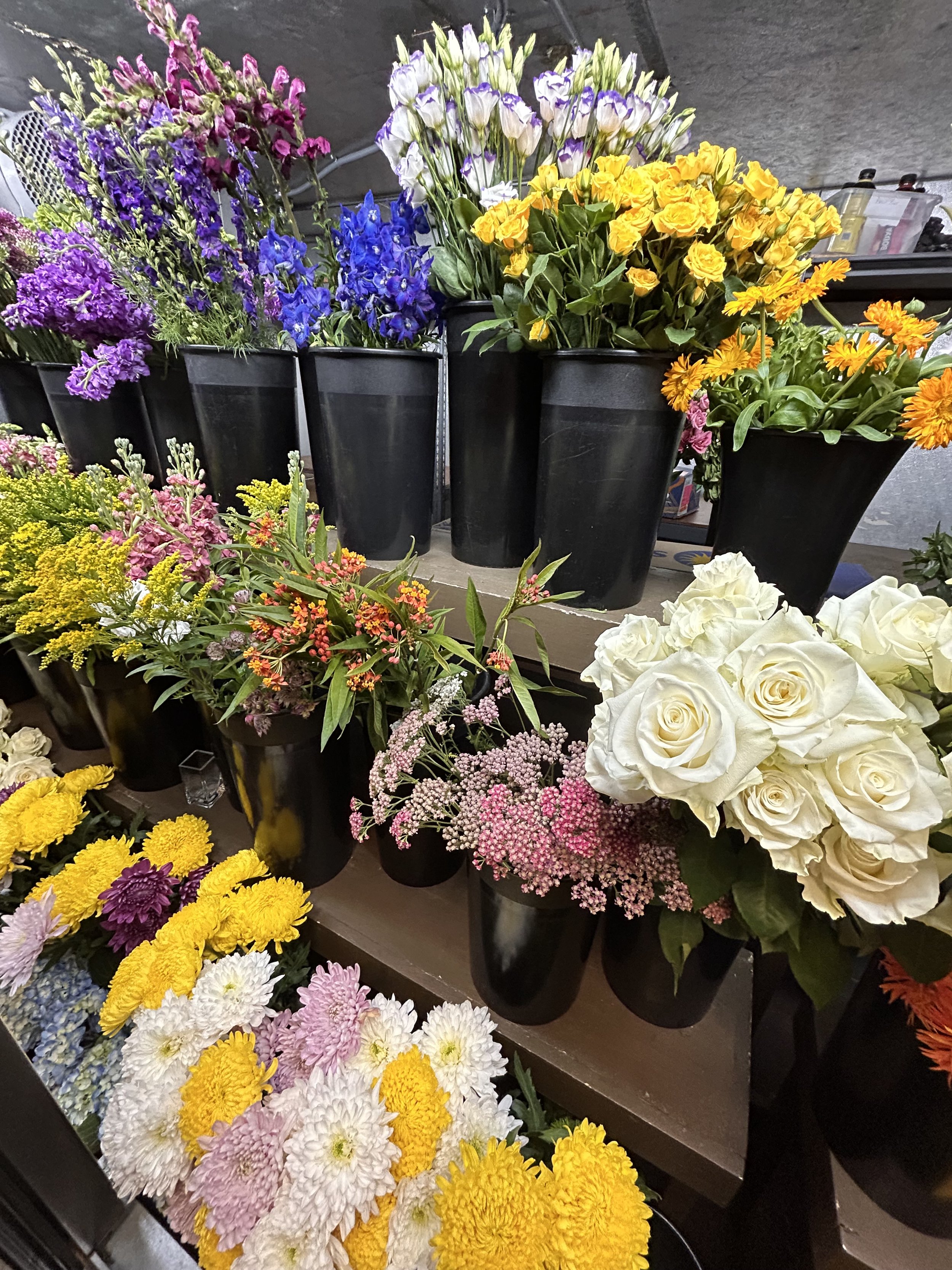 Various colorful flowers in black pots on shelves, including white roses, yellow daisies, pink and purple statice, blue delphinium, and other mixed flowers in a flower shop.