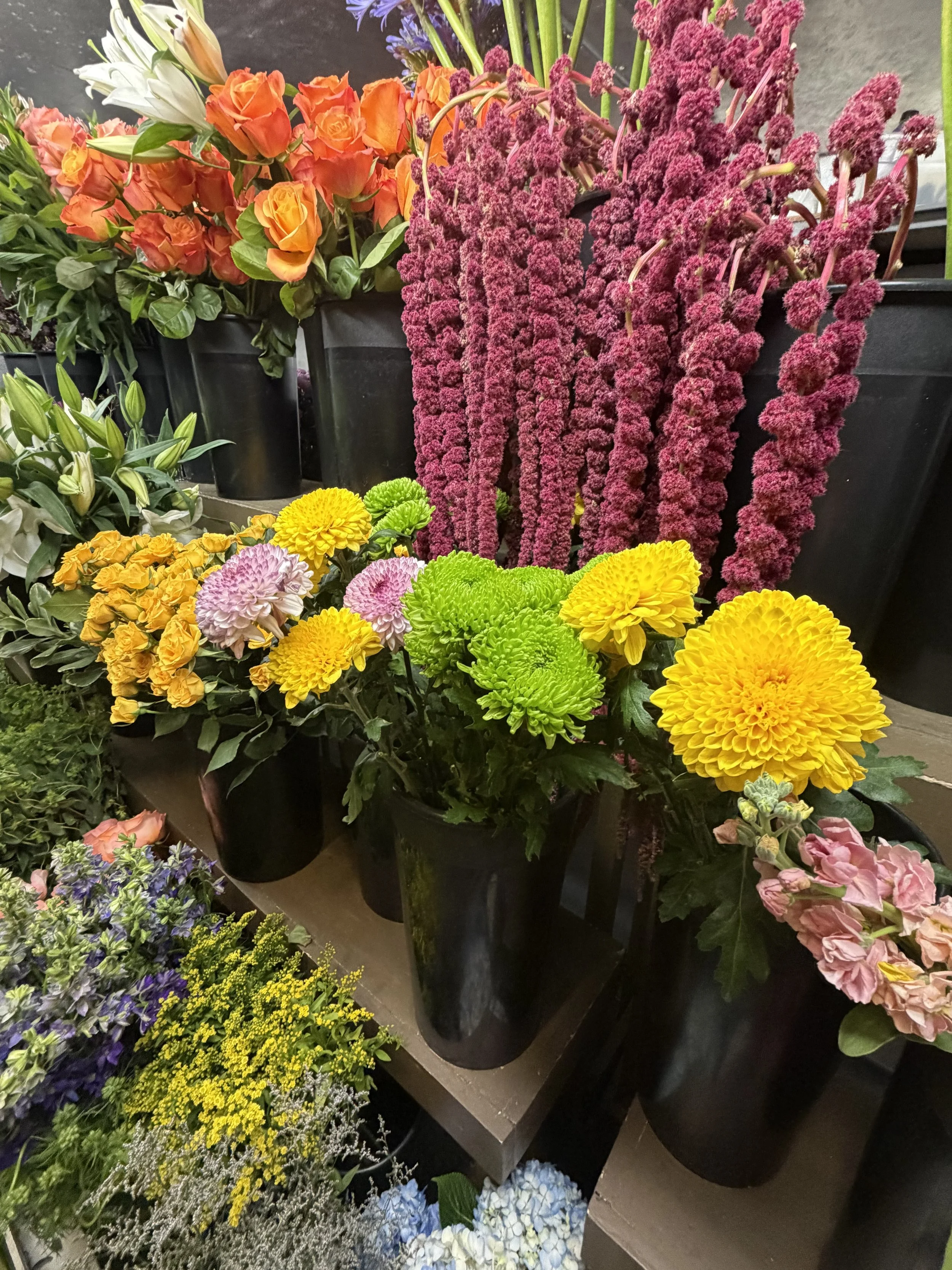 Various colorful flowers, including orange roses, white lilies, pink chrysanthemums, yellow chrysanthemums, purple asters, blue hydrangeas, and other mixed flowers, arranged in black vases on display.