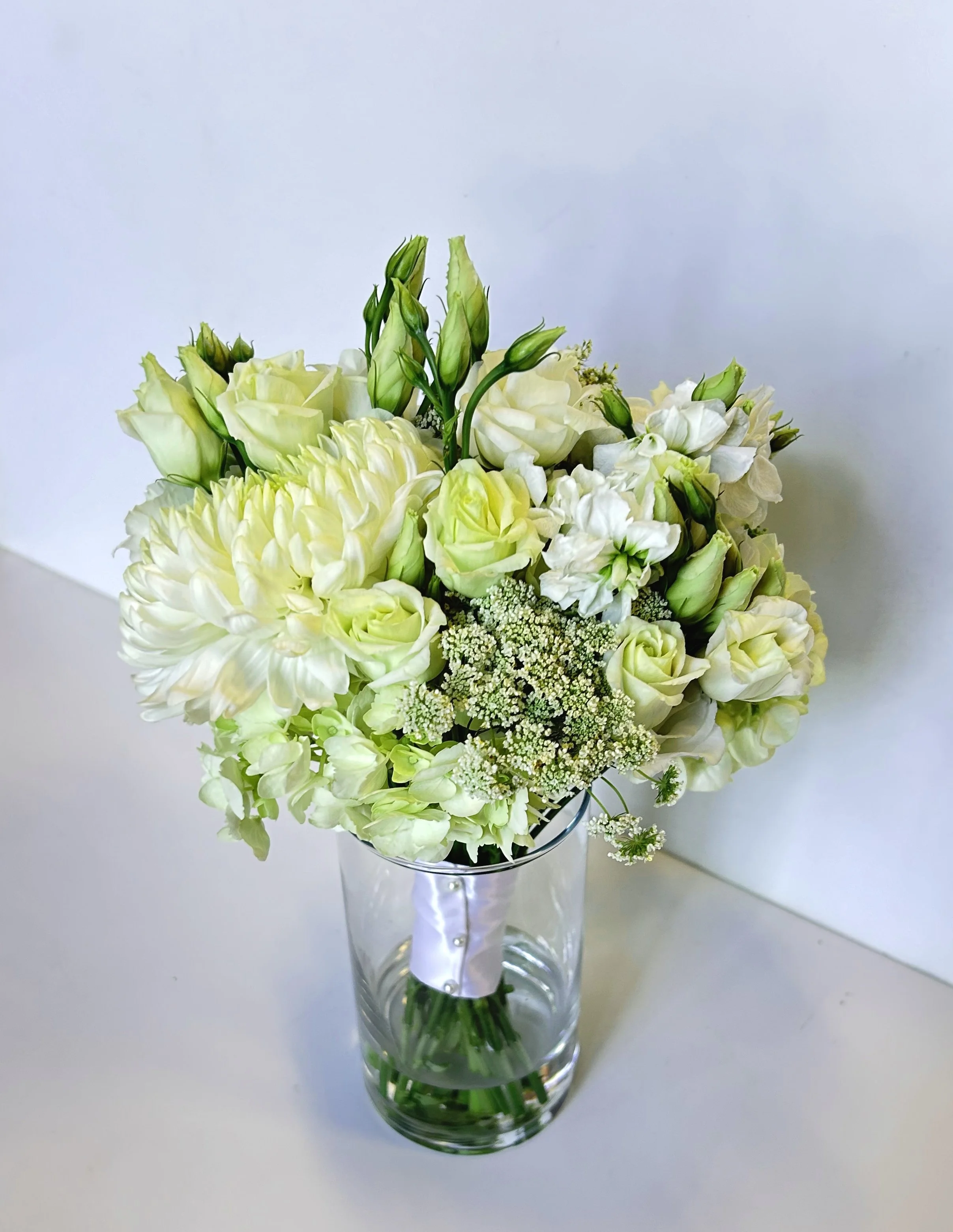 A Nosegay or prom bouquet of white flowers in a clear glass vase on a white surface.