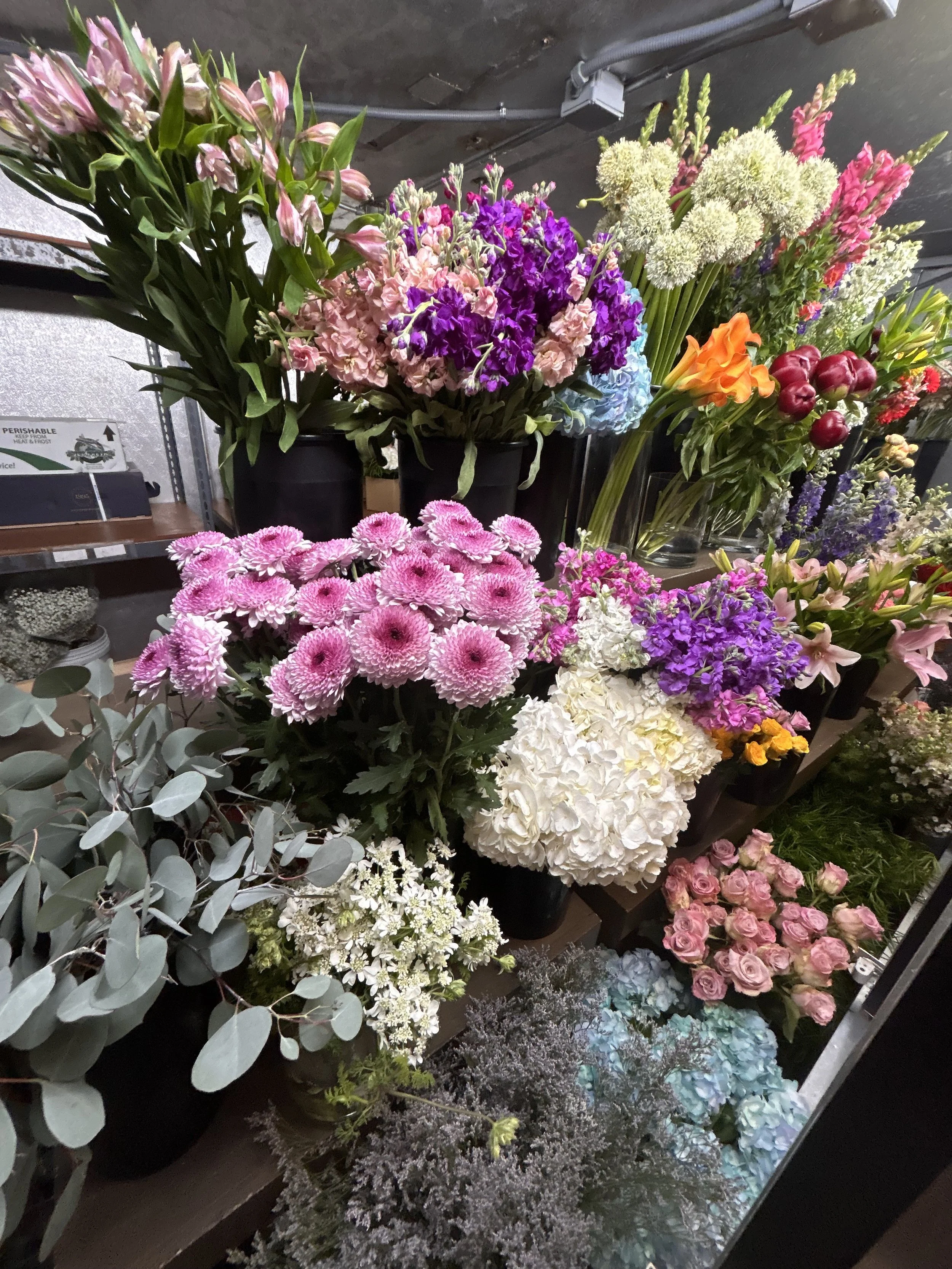 A display of various colorful flowers in black pots on shelves in a floral shop, including pink, purple, white, orange, and red flowers with greenery.