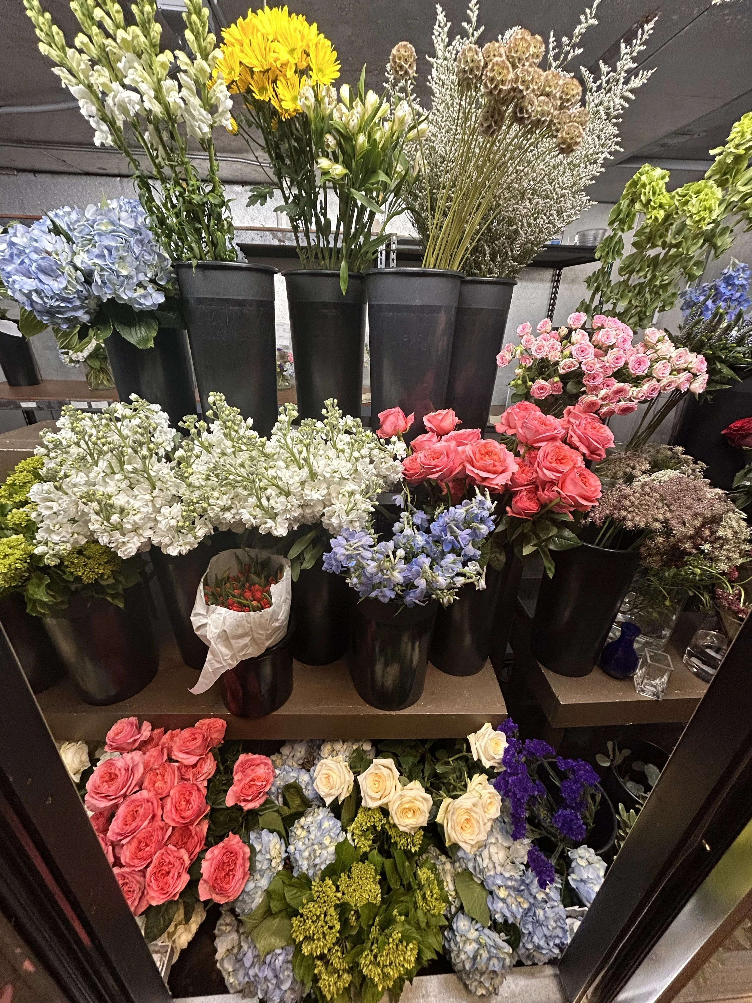 Various colorful flowers in black pots and wrap, including hydrangeas, roses, lisianthus, and other flowers, arranged on shelves and the floor in a flower shop.