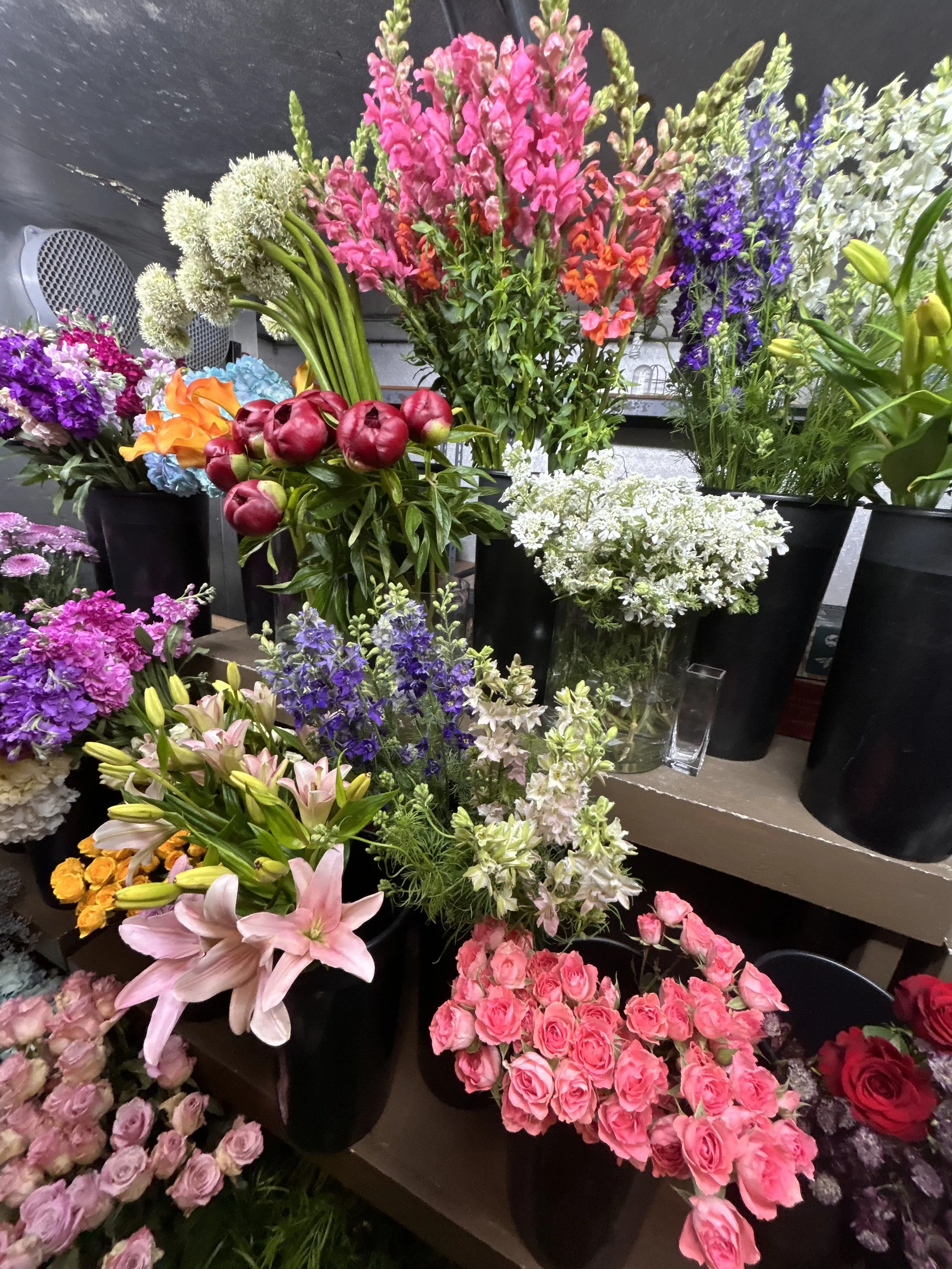 Various colorful flowers including pink lilies, roses, purple, white, and red flowers in black pots on display.