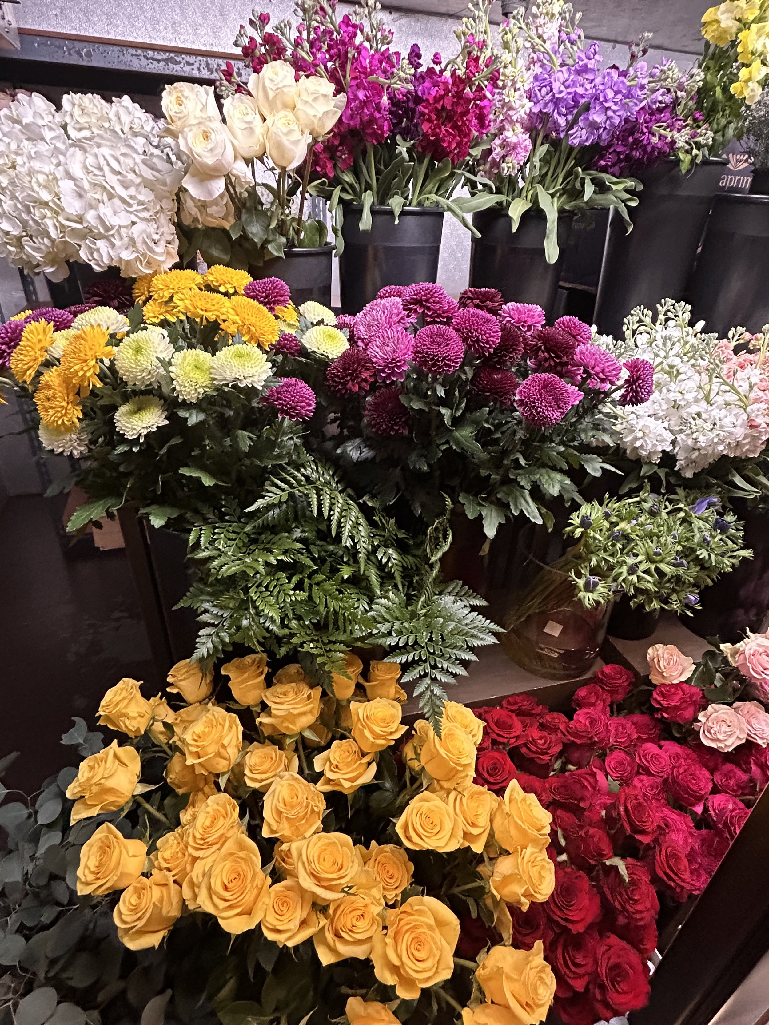 A variety of colorful flowers including yellow roses, white daisies, pink and purple chrysanthemums, white hydrangeas, and pink roses arranged in black buckets.