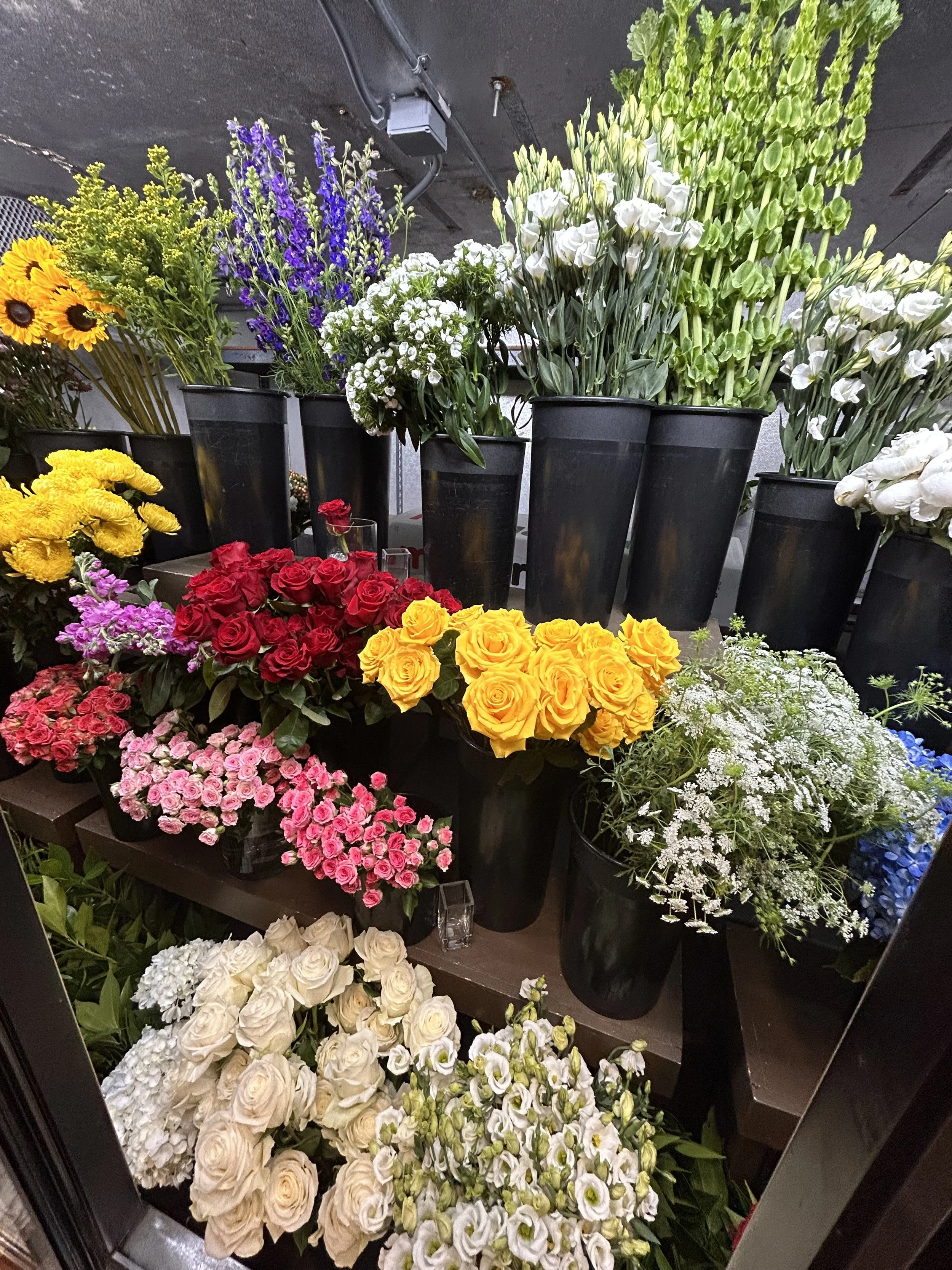 A display of various colorful flowers arranged in black pots, including sunflowers, purple, white, red, yellow, and pink flowers, set in an indoor market or store with a dark ceiling.