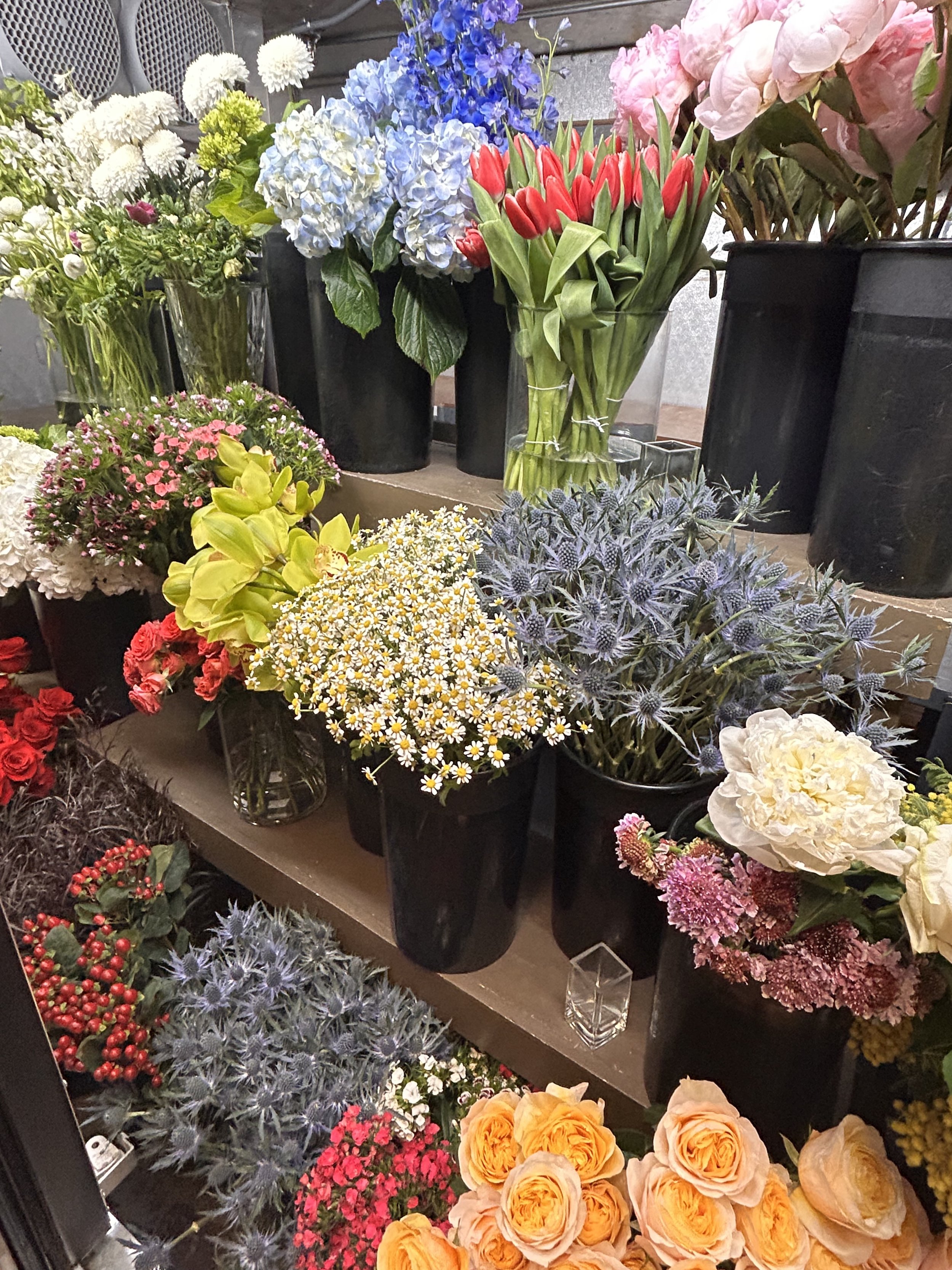 Arrange of colorful flowers including hydrangeas, tulips, roses, daisies, and other blooms in various vases and pots, displayed on a wooden shelf.