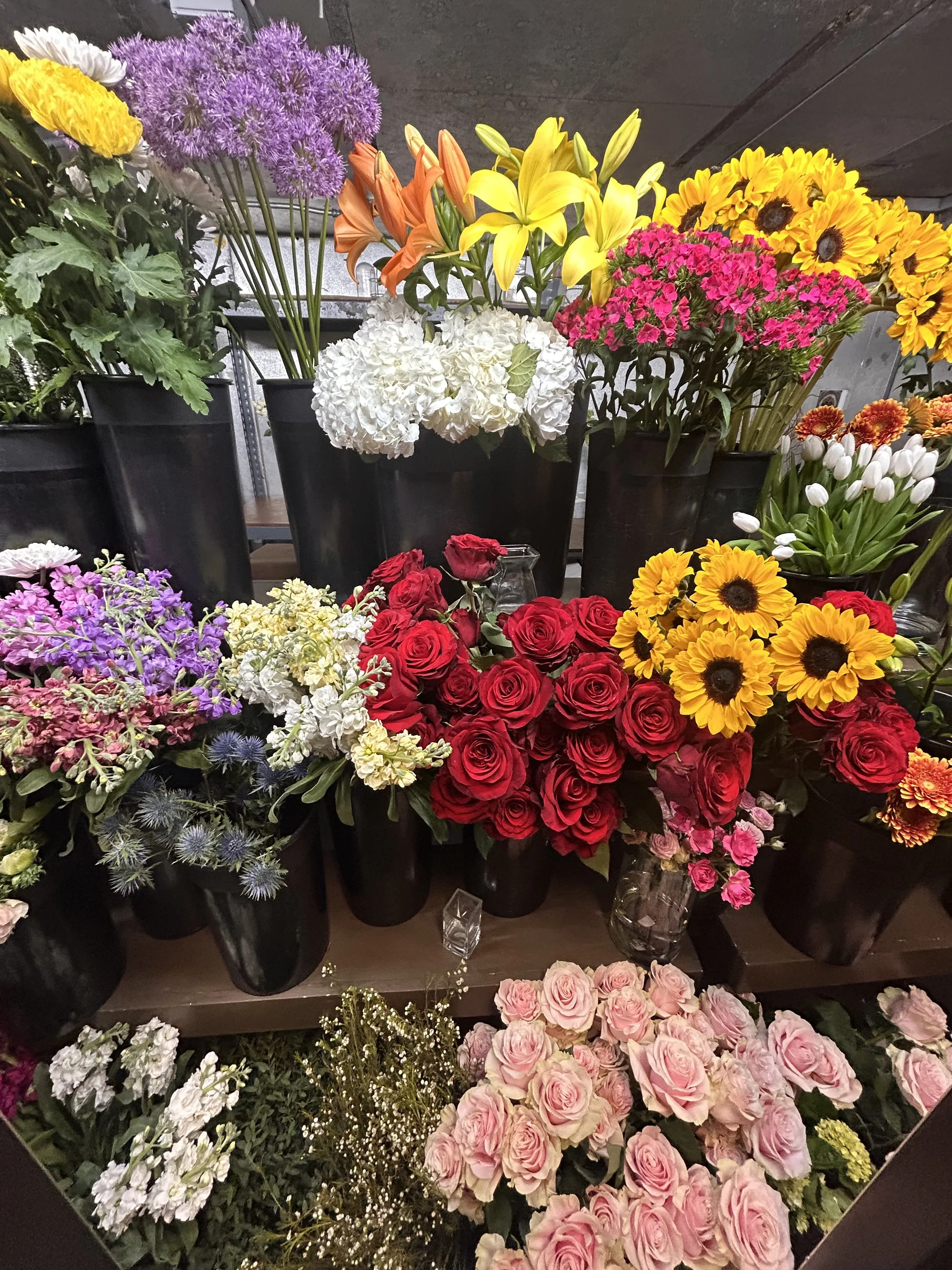 Various colorful flowers, including roses, sunflowers, tulips, and lilies, arranged in black pots and vases on a display shelf.