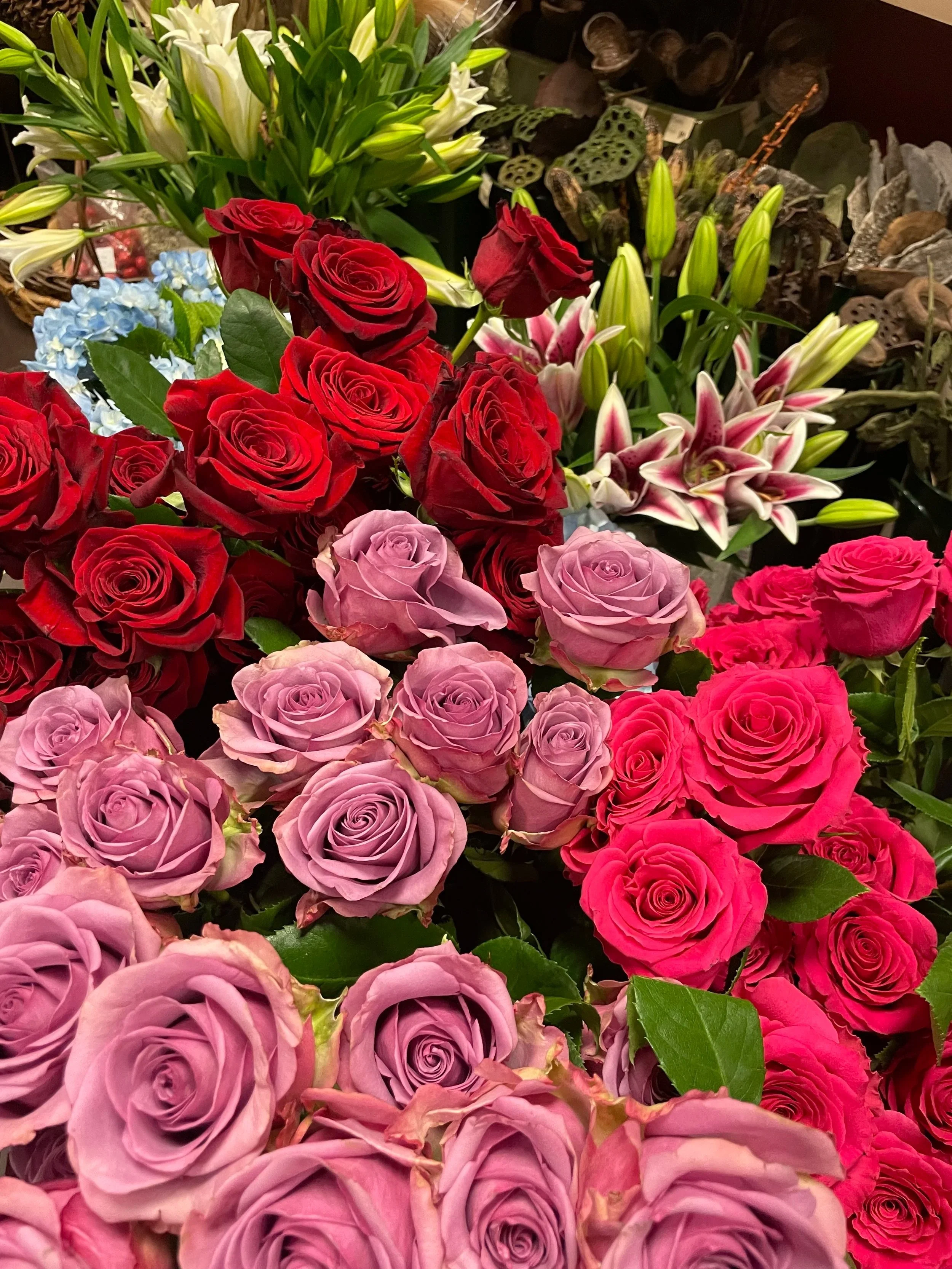 A display of various colorful flowers including red roses, hot pink roses, lavender roses, white lilies, and pink lilies displayed together.