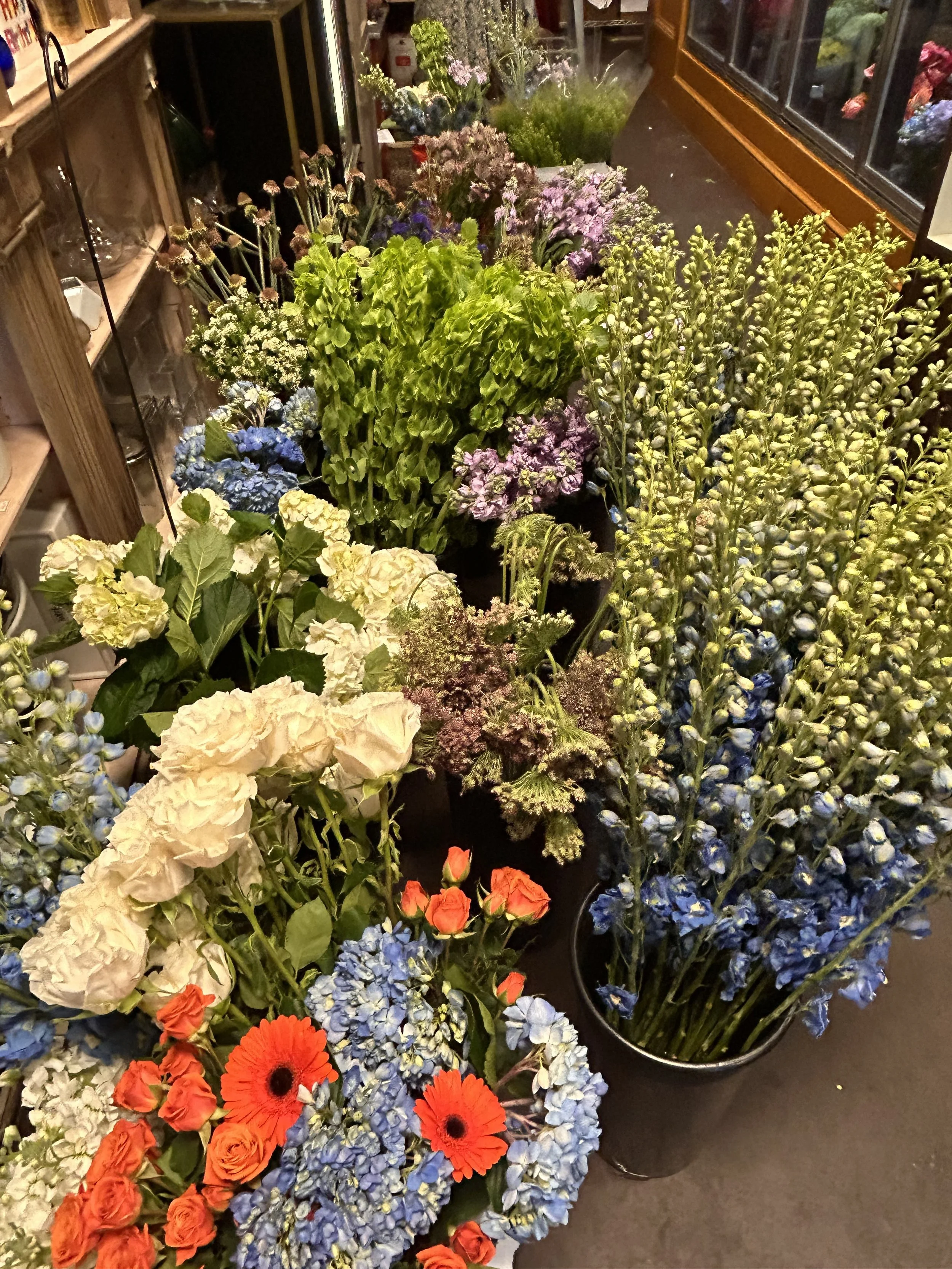 Various colorful flowers including red and orange gerbera daisies, white hydrangeas, purple hydrangeas, and yellow flowers in vases and pots in a store.
