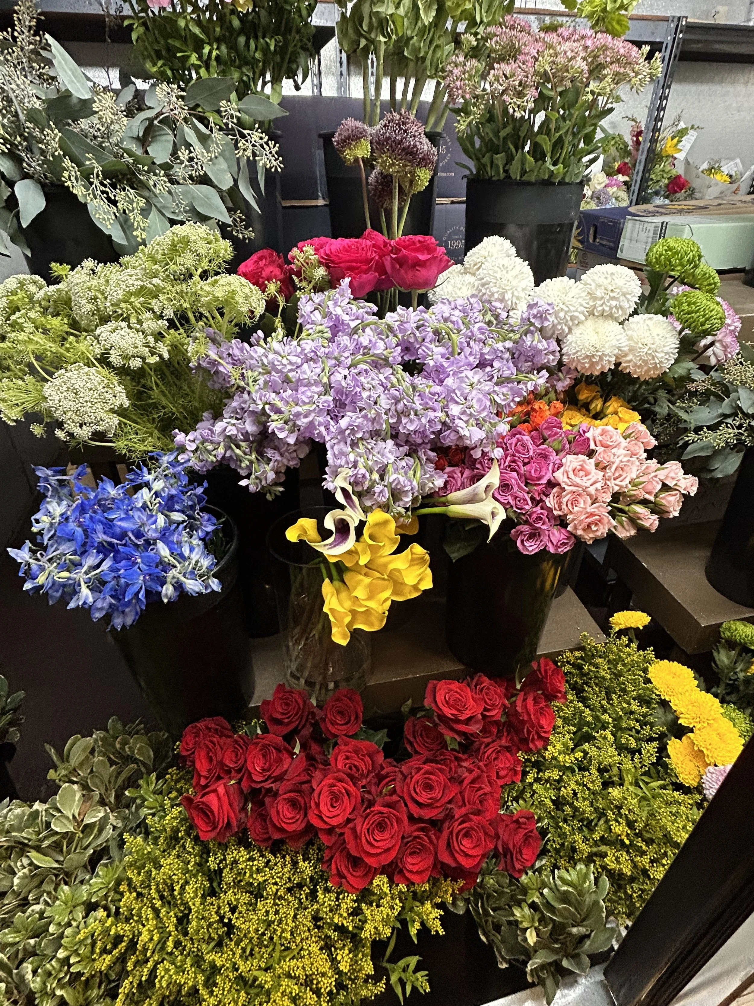 An assortment of colorful flowers including roses, lilies, and various fresh cut blooms arranged in vases on a table, with some greenery in the background.