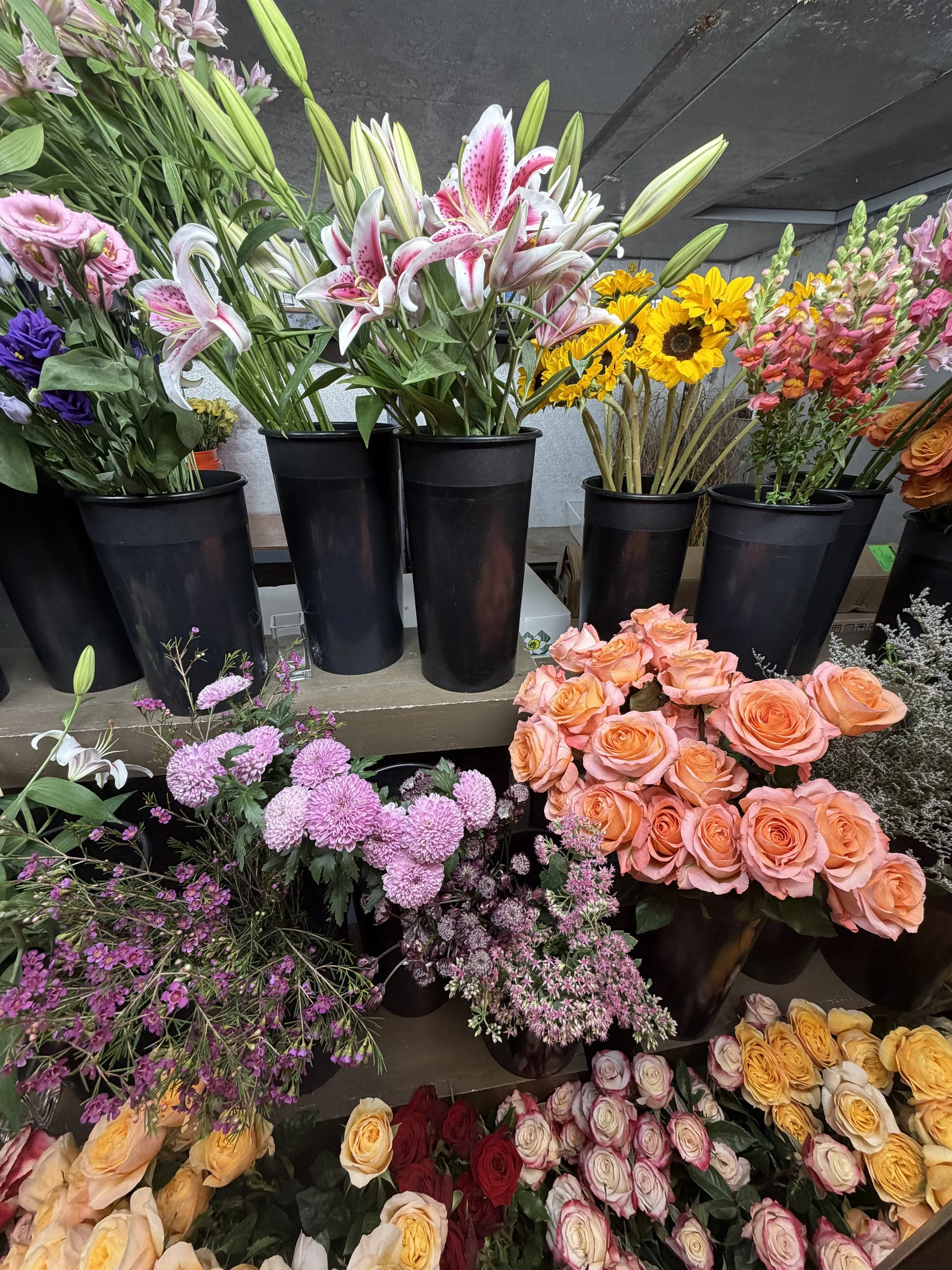 Various colorful flowers including pink lilies, yellow sunflowers, peach and pink roses, and purple and white flowers arranged in black pots at a flower shop.