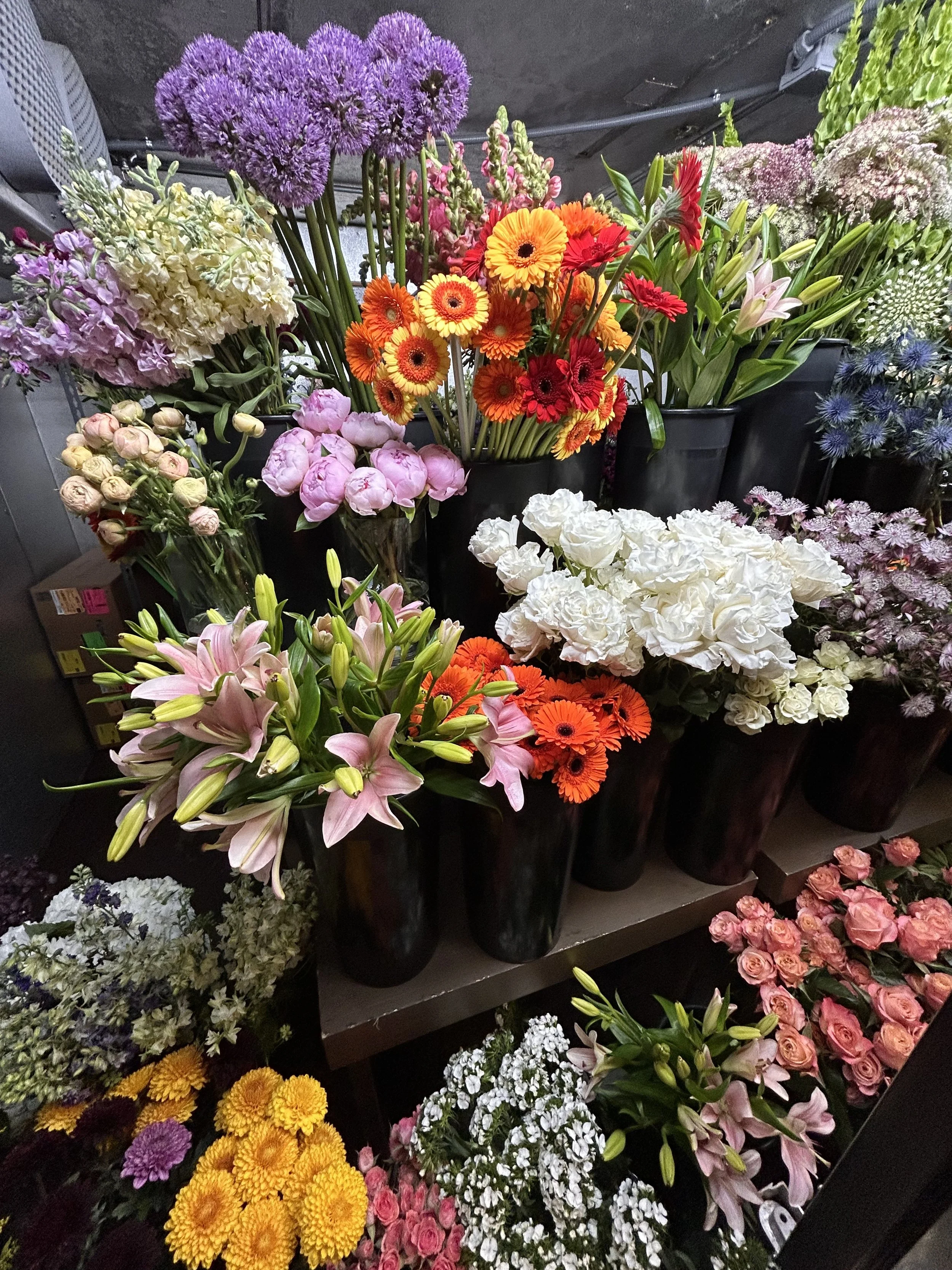 A variety of colorful flowers on display, including pink roses, yellow and orange chrysanthemums, purple alliums, pink lilies, white hydrangeas, and other mixed flowers arranged in black pots and vases.
