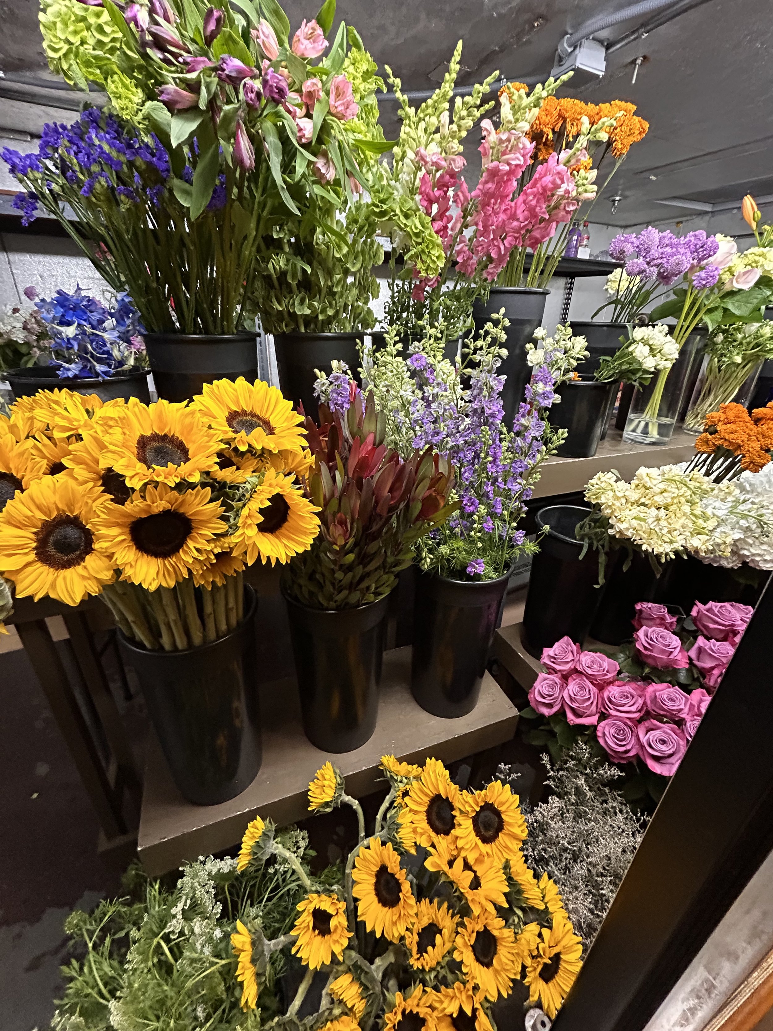 A variety of colorful flowers, including sunflowers, pink roses, purple, orange, and white flowers, arranged in black vases on a display shelf in a flower shop.