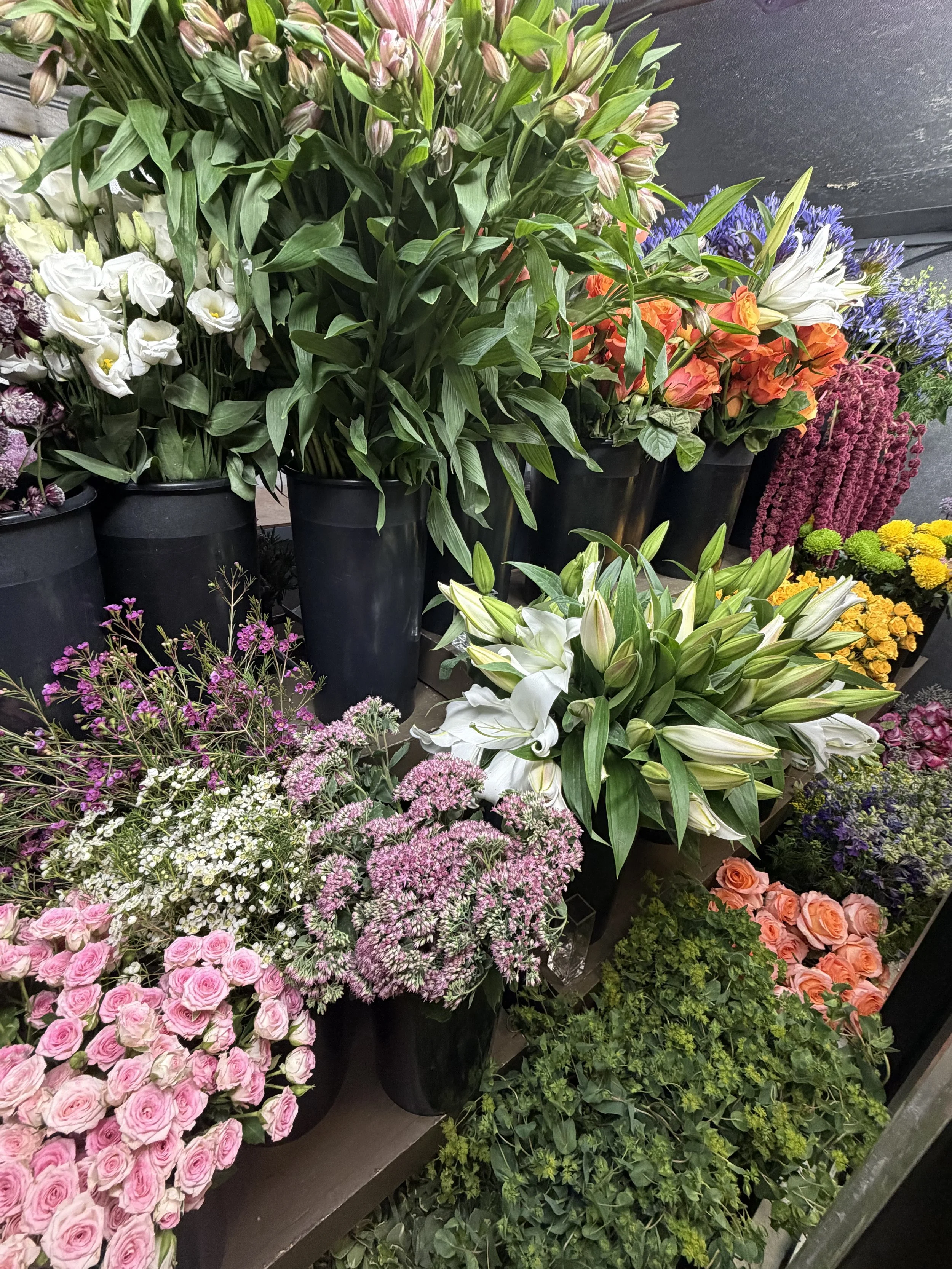 A display of various colorful flowers in black buckets, including pink roses, white lilies, purple statice, orange and white lilies, and other assorted blooms.