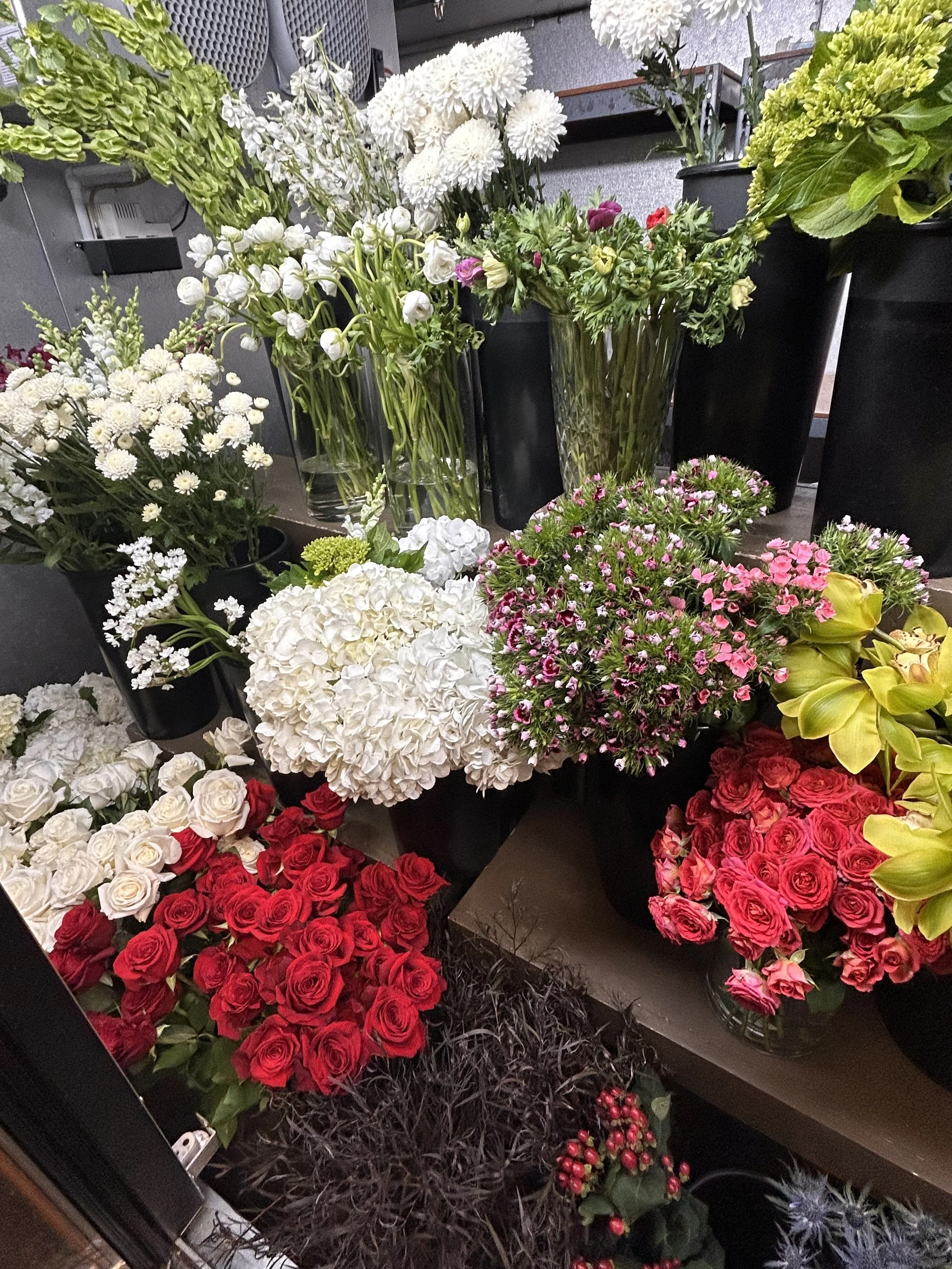 A variety of flowers including red roses, white hydrangeas, pink daisies, and white chrysanthemums displayed in black vases and containers.