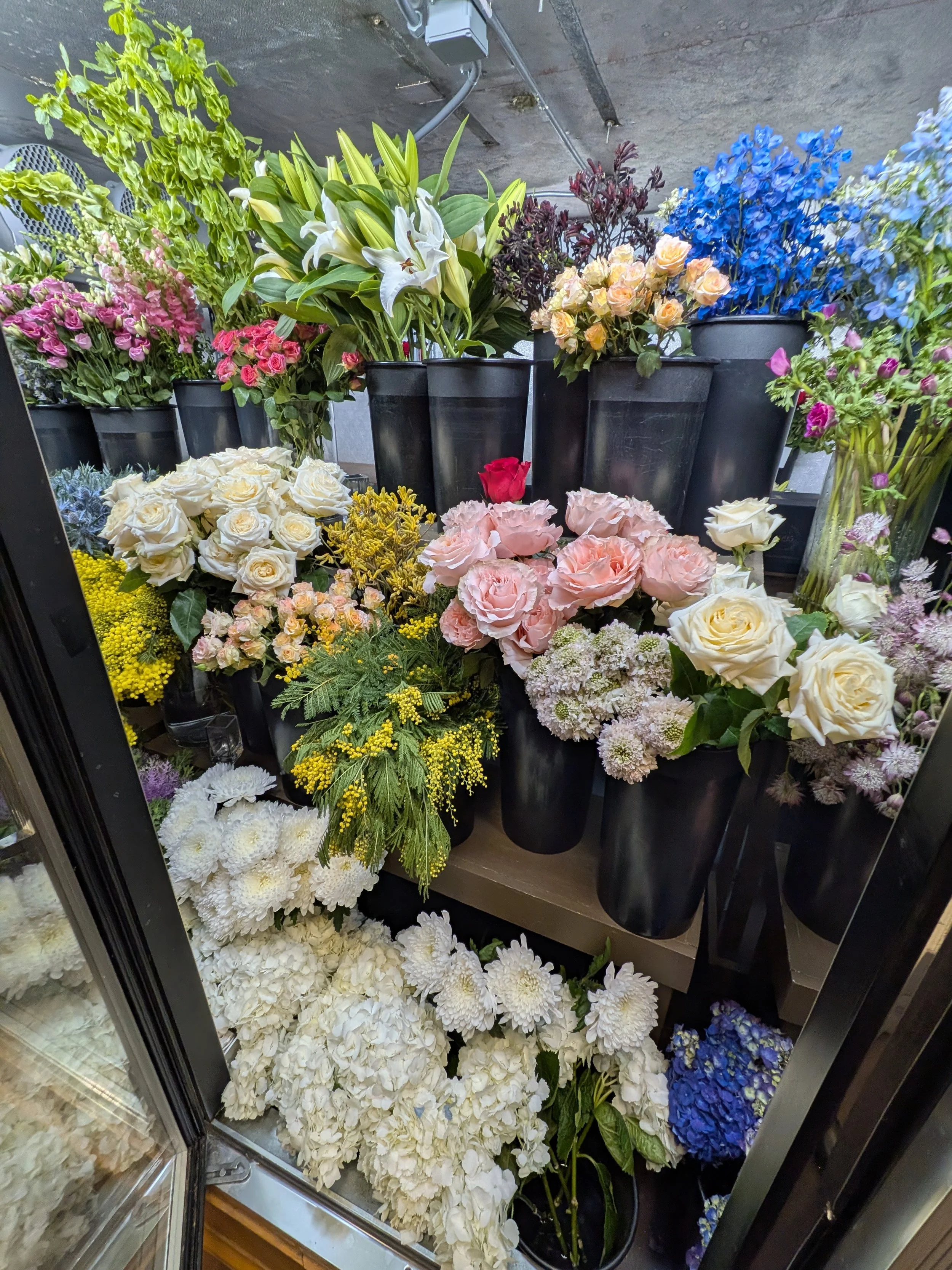 A display of various colorful flowers in black pots, including white lilies, pink roses, yellow daisies, white hydrangeas, blue and purple flowers, and other mixed blooms, arranged on shelves and the floor.