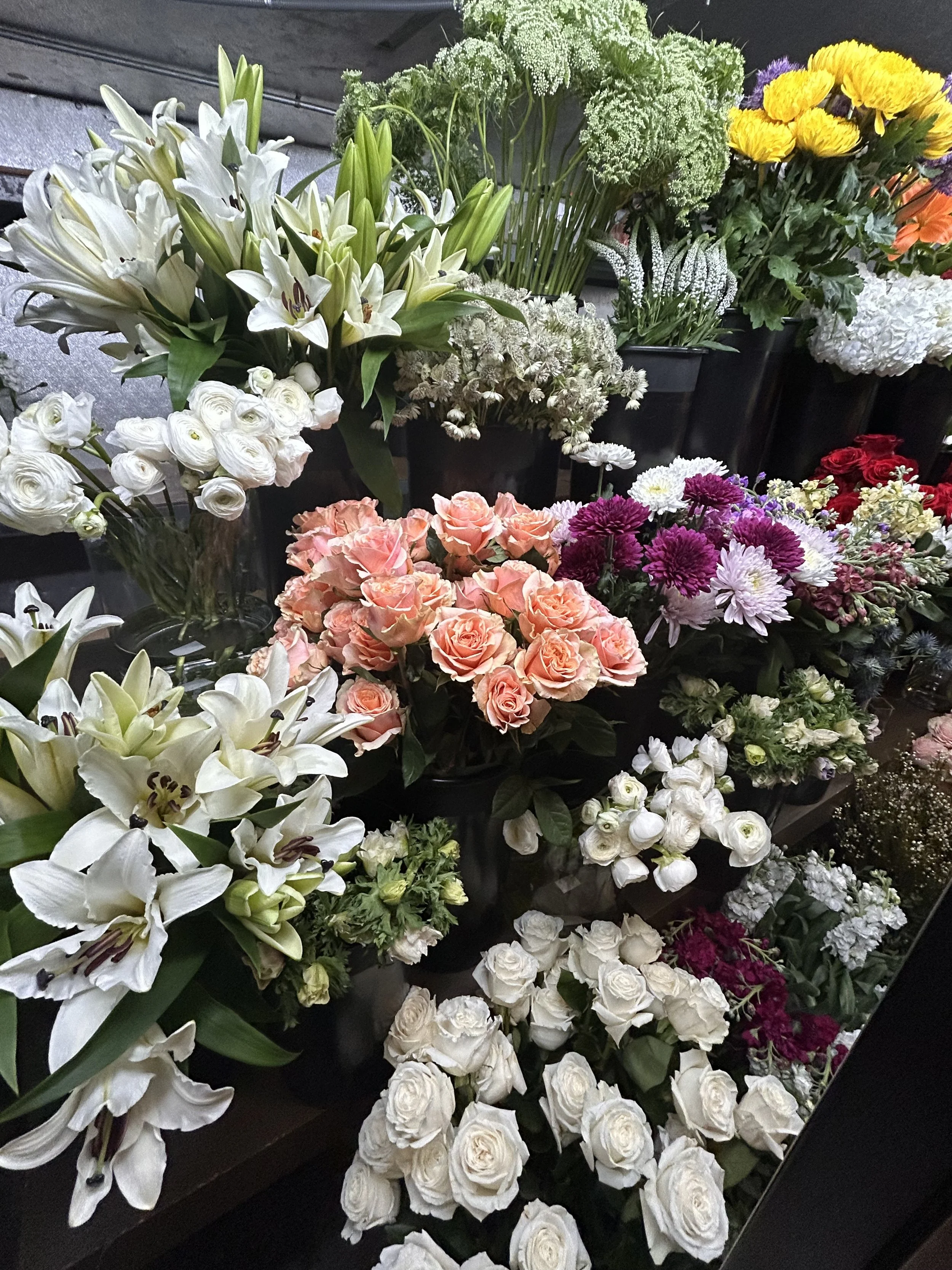 Various colorful flowers including white lilies, white roses, pink roses, purple chrysanthemums, and yellow blooms displayed in black pots.