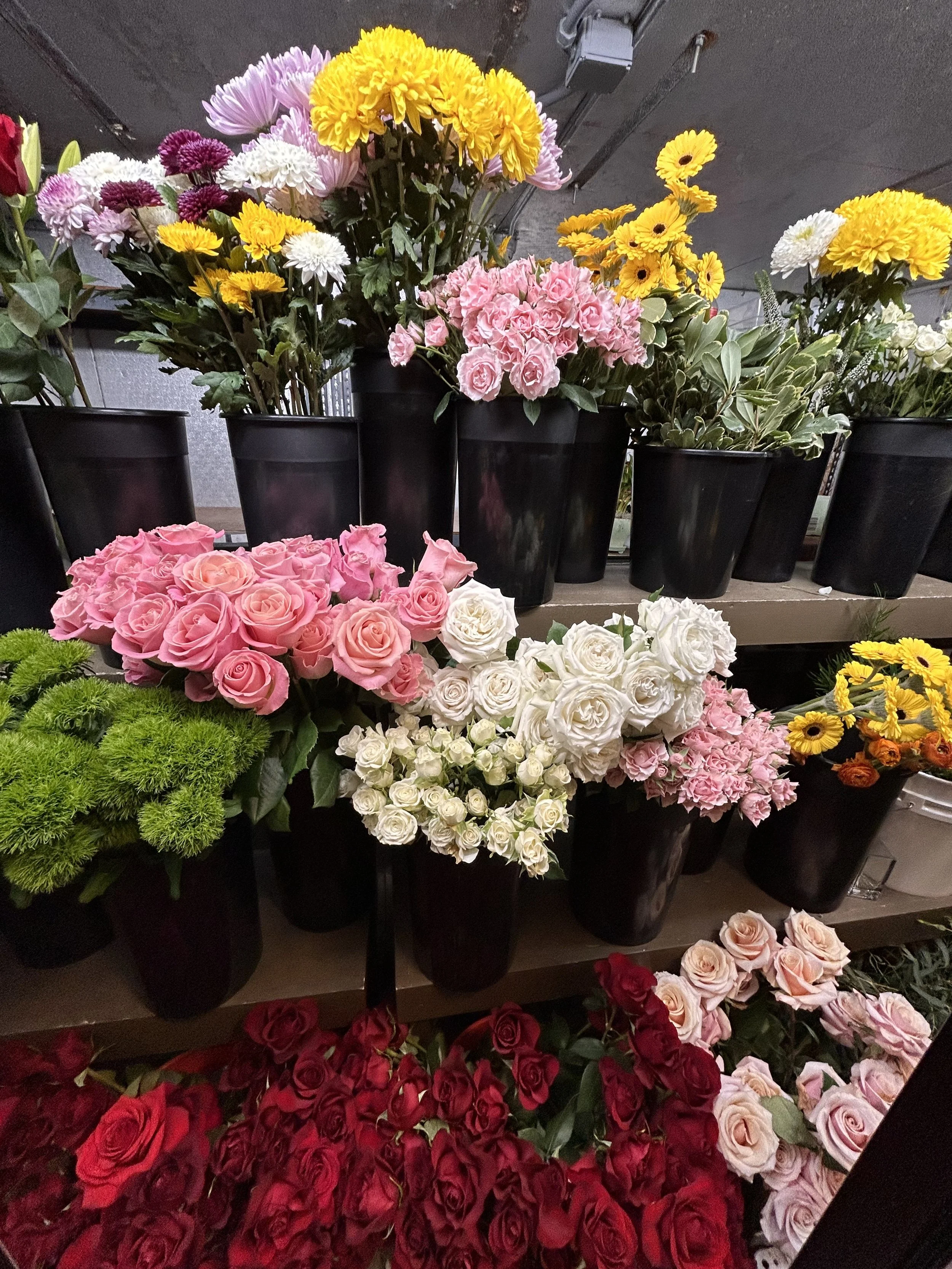 Assorted colorful flowers in black pots, including pink roses, white roses, yellow daisies, pink chrysanthemums, and red roses, arranged on shelves.