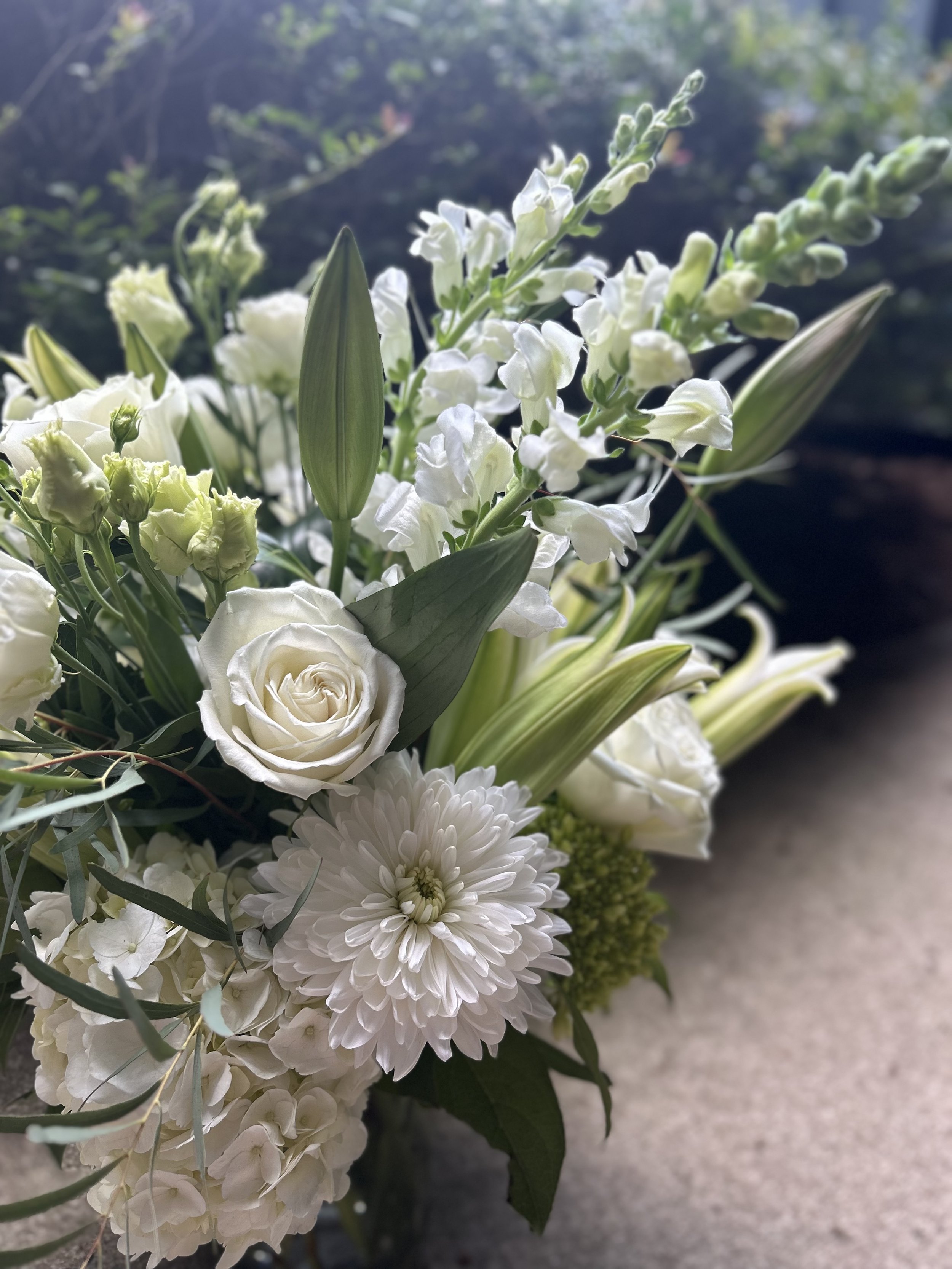 Close-up of a white floral bouquet with roses, lilies, and other white flowers, set against a blurry outdoors background.
