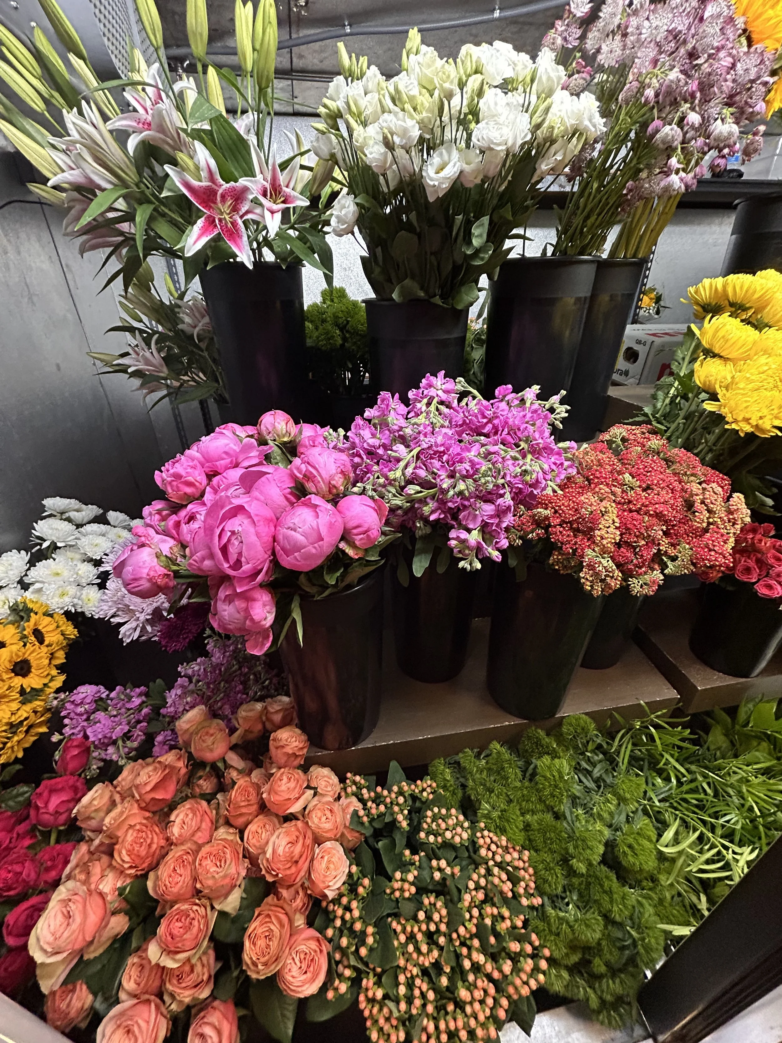 A collection of colorful flowers arranged in black vases and containers, including lilies, roses, peonies, milkweed, daisies, and other mixed floral varieties.