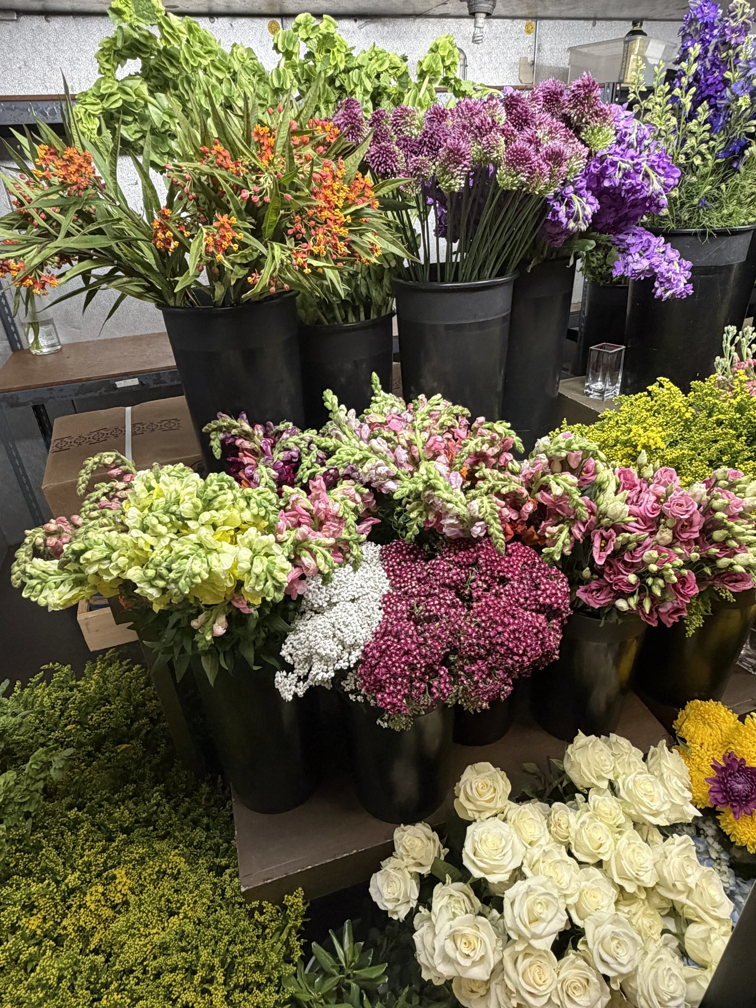 Various colorful flowers and plants arranged in black pots and displays at a flower shop or market, including purple, pink, white, and yellow blossoms.