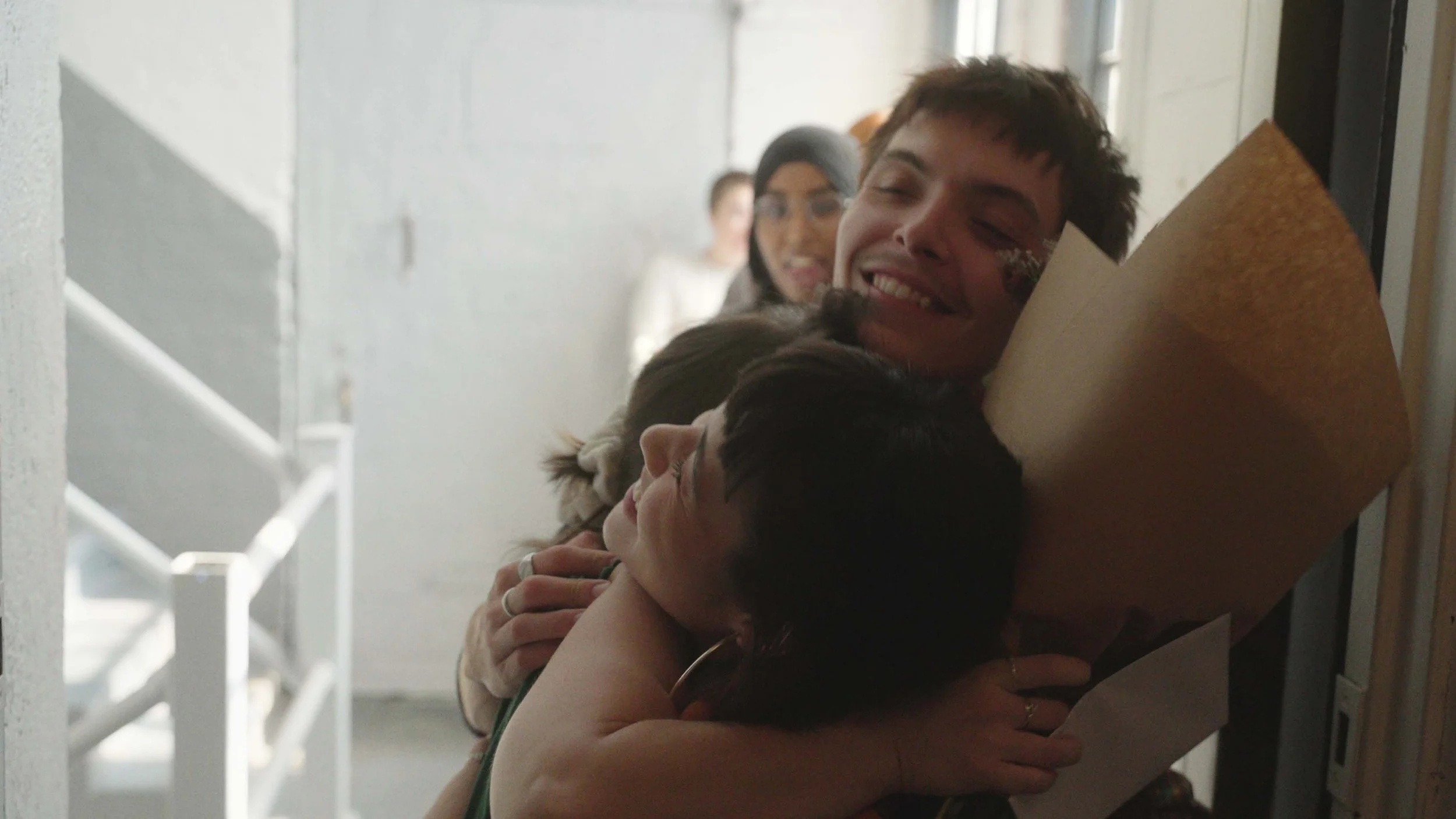 Two people embracing in a hug, with surrounding individuals smiling and observing in the background, in an indoor setting.