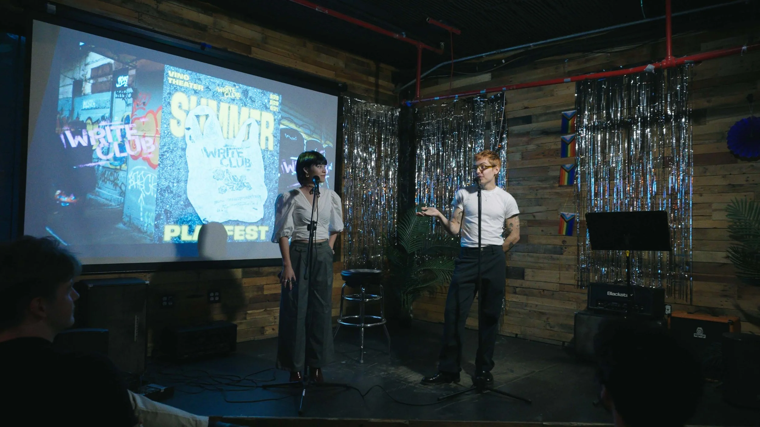 Two people stand on a stage with microphones, engaged in a discussion, in front of a projection displaying "SQUAR" and "WIRED CLUB" with a large illustration of a bag, in a decorated venue with shimmering curtains and South African flag decorations.