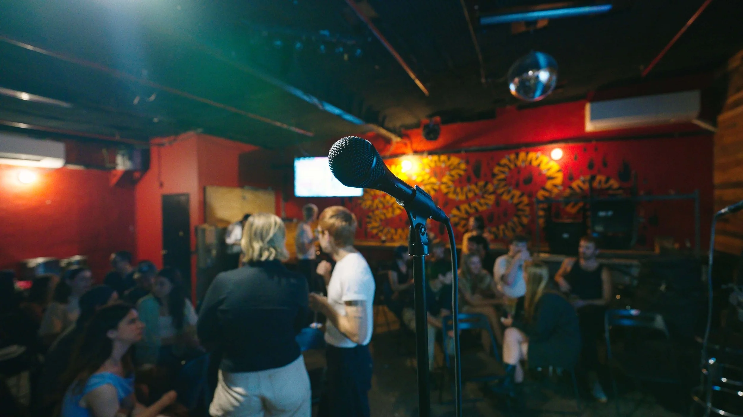 Microphone on a stand in the foreground of a dimly lit comedy club or open mic venue, with people sitting and socializing in the background, and colorful wall art behind them.