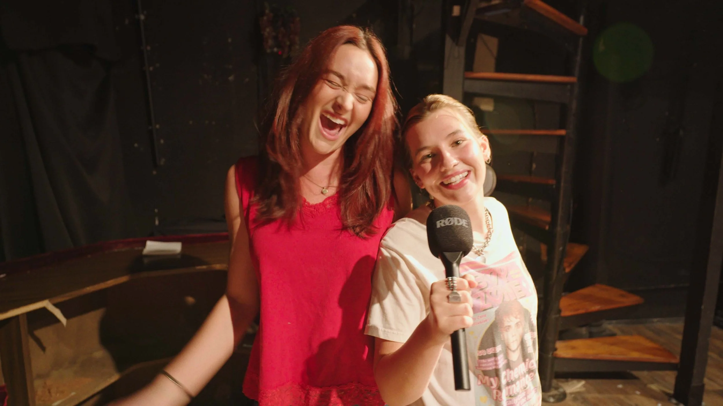 Two women smiling and laughing, one holding a microphone, in a dimly lit indoor space with black backdrop and wooden shelves.