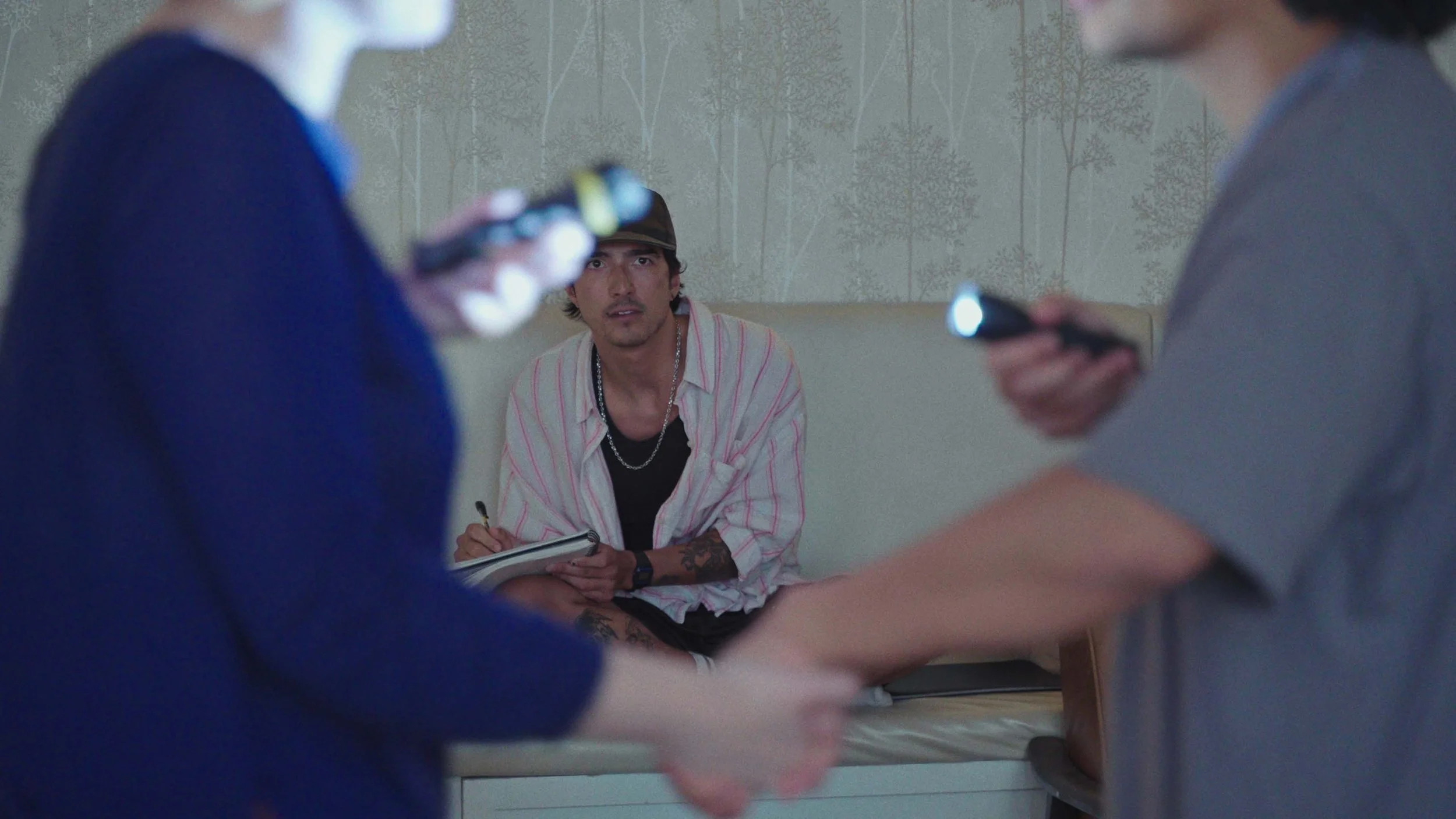 A man wearing a striped shirt and a hat is sitting on a bed, holding a notebook and pen, looking surprised at two women in front of him who are holding flashlights.