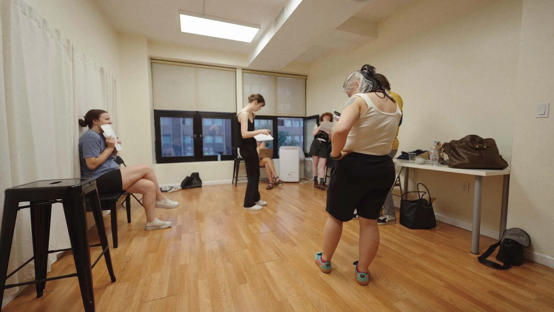 Group of women in a room with wooden floors, some standing, some sitting, engaged in various activities, with a large window and a skylight overhead.