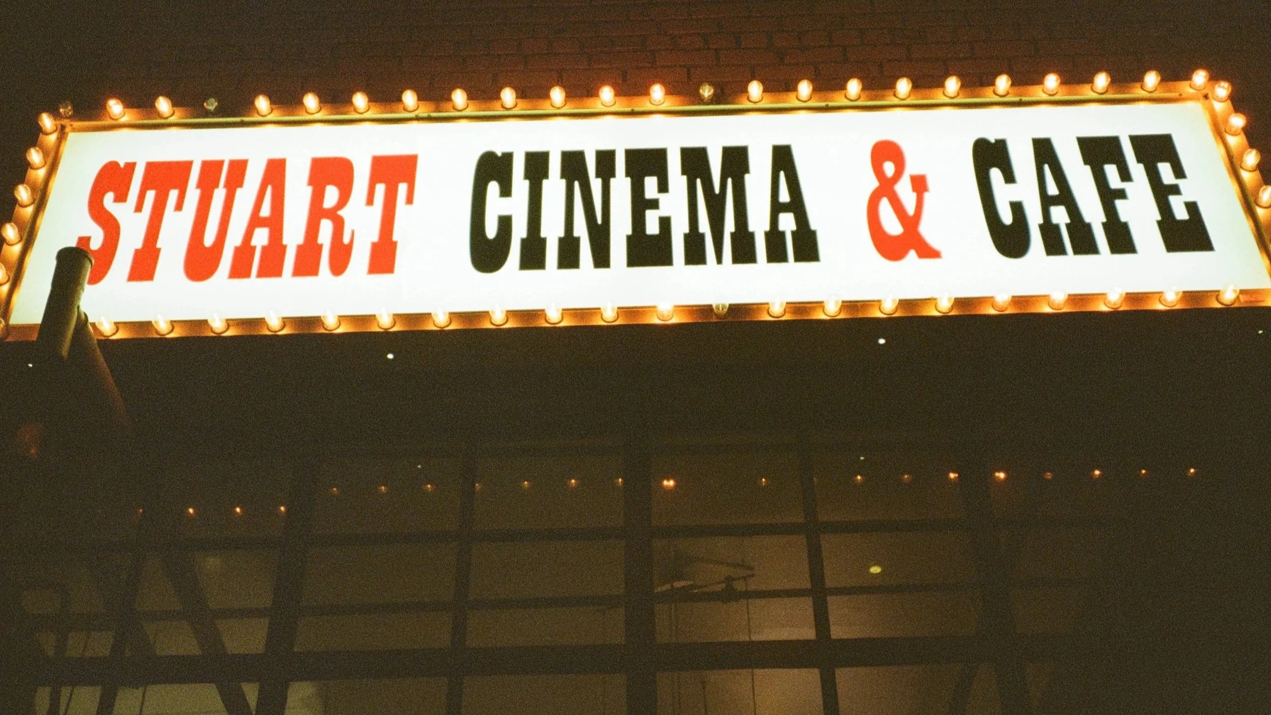 Night view of a bright marquee sign reading 'Stuart Cinema & Cafe' with lights surrounding the sign, black letters for 'Cinema & Cafe', and red letters for 'Stuart.'