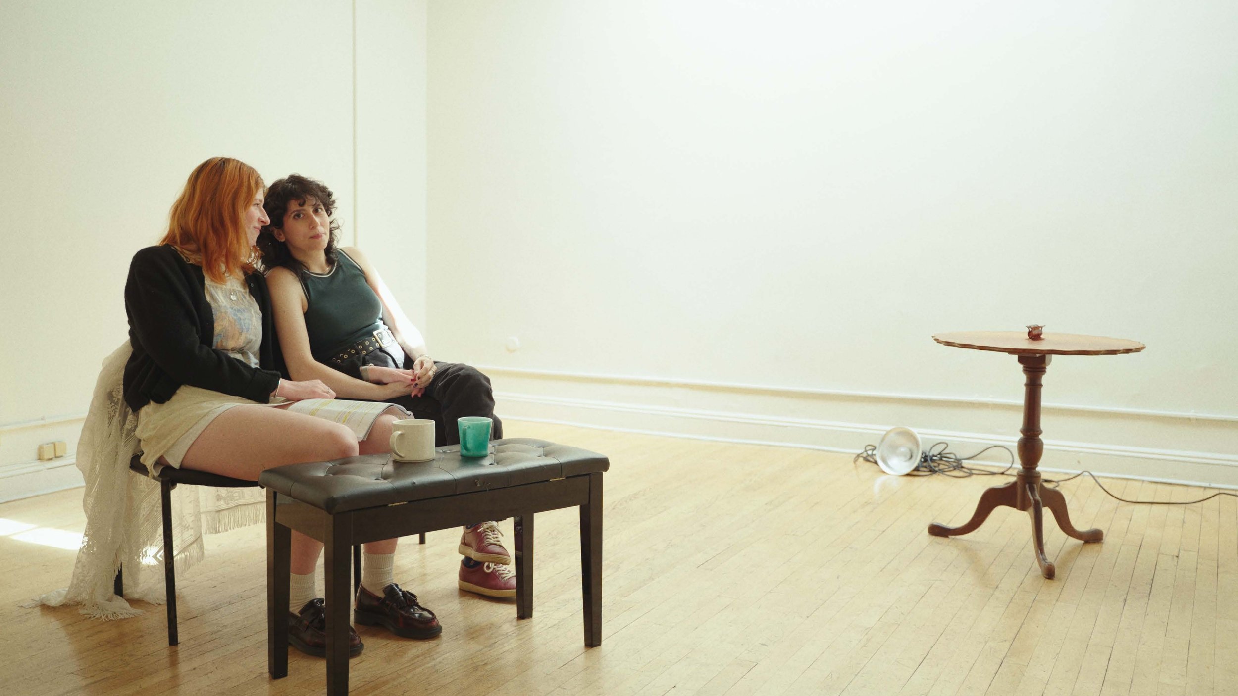 Two women sitting on a bench in a sparse room with minimal furniture, a small table to the right, and some scattered cords and a light fixture on the floor.