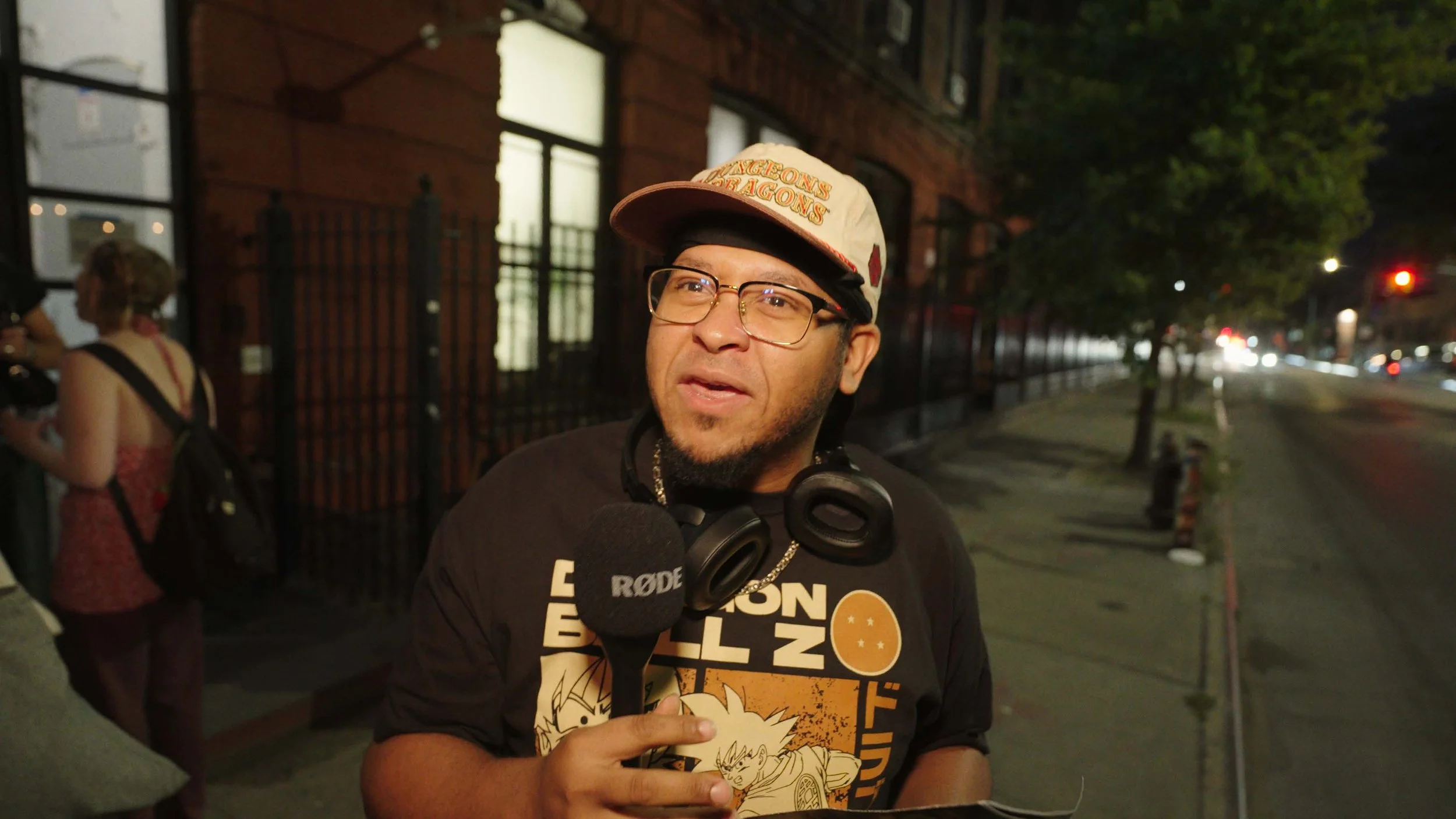 A man holding a microphone labeled 'RODE' speaking outdoors at night, wearing glasses, a beige cap with 'DRAGONS' written on it, and a black T-shirt with anime graphics. He has headphones around his neck and is pointing with his finger. In the background, there is a woman with curly hair, a backpack, and a red dress, standing near a brick building with large windows. The street is lined with trees and streetlights.