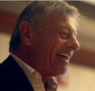 Profile of a smiling middle-aged man with short gray hair, wearing a checked shirt and jacket.
