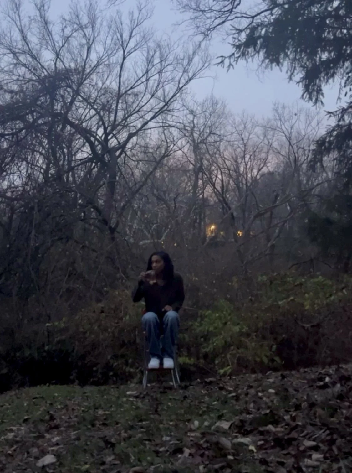 A woman sitting on a chair outdoors among leafless trees during dusk, drinking from a glass.