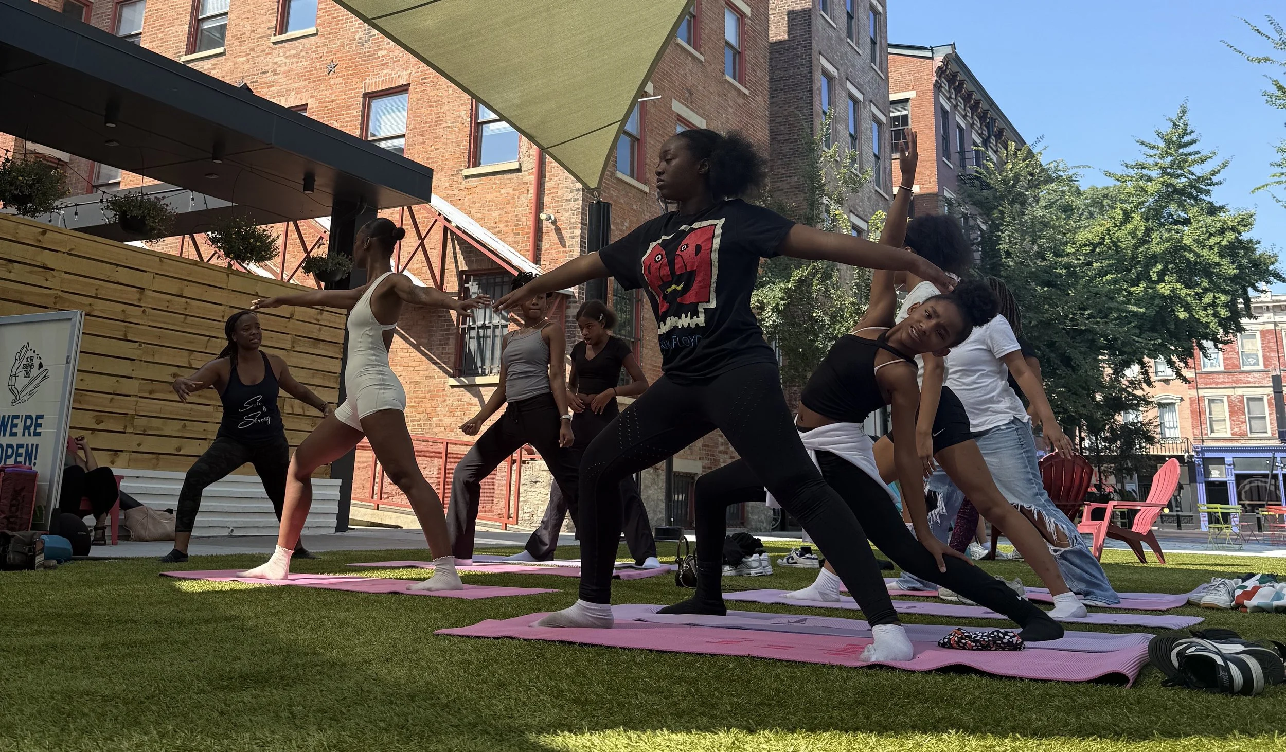 Group of women participating in outdoor yoga class on pink mats, performing stretching poses in urban park with brick buildings and trees in background.