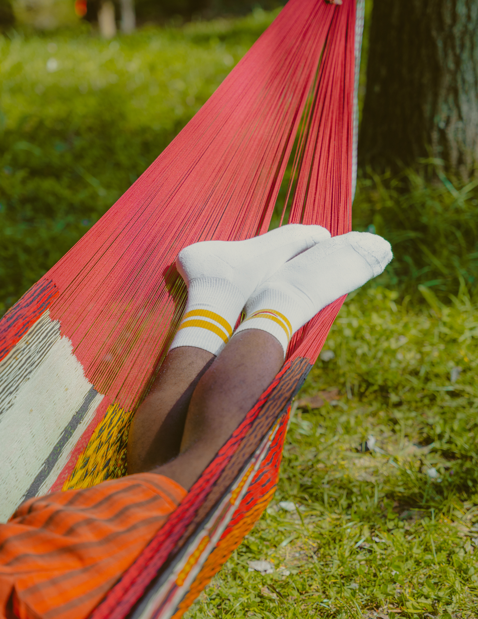 Person lying on a red hammock wearing white socks with yellow stripes, orange and black checkered shorts, and a green shirt, in an outdoor grassy area next to a tree.
