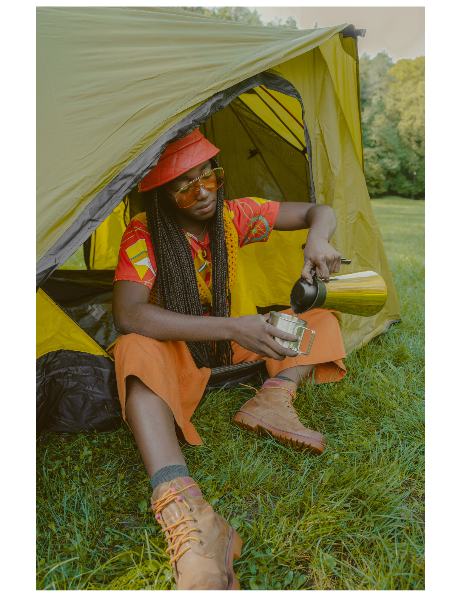 A person sitting on the grass outside a yellow camping tent, pouring coffee from a thermal carafe into a mug, wearing a red hat, orange boots, and a colorful shirt.