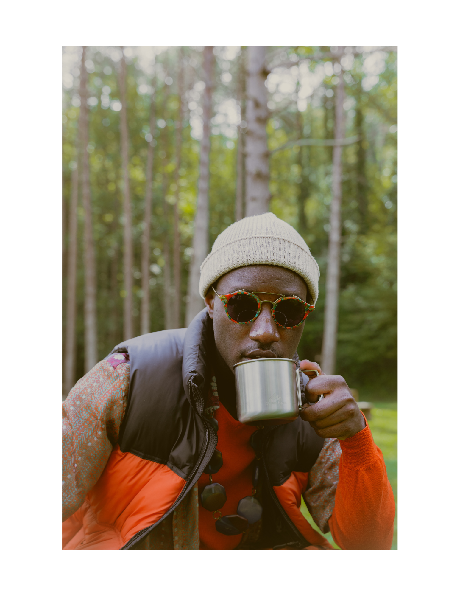 A person wearing a beige knit hat, patterned sunglasses, and an orange and black vest, holds a metallic mug while outdoors in a forest with tall green trees.